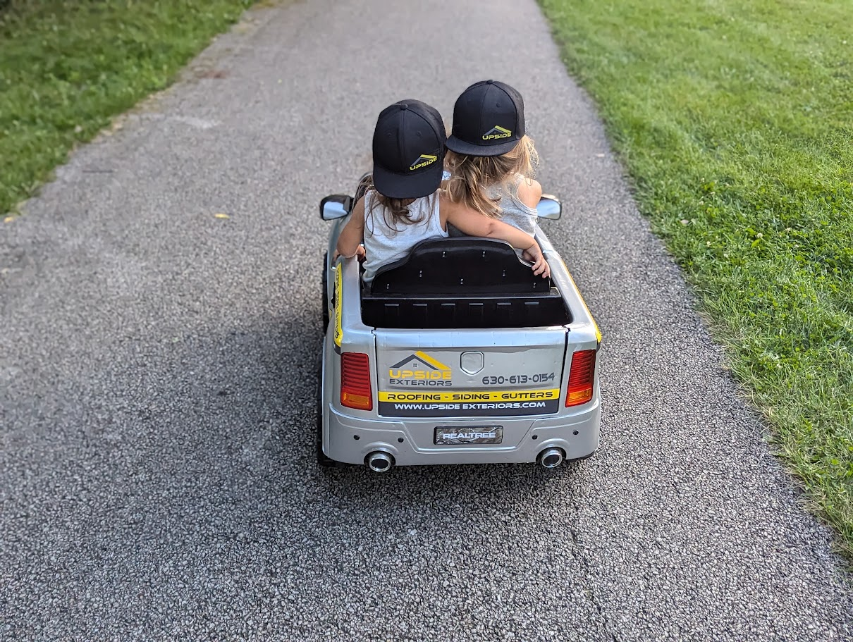 Two daughters riding together in a miniature Upside Exteriors truck on a paved path.