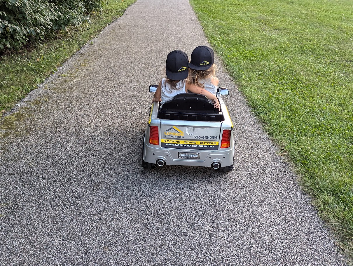 Two daughters driving a miniature Upside Exteriors truck down a paved path.