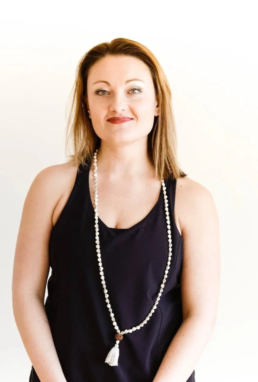 A woman with shoulder-length auburn hair wearing a black sleeveless top and a long beaded necklace with a tassel, standing against a plain white background.