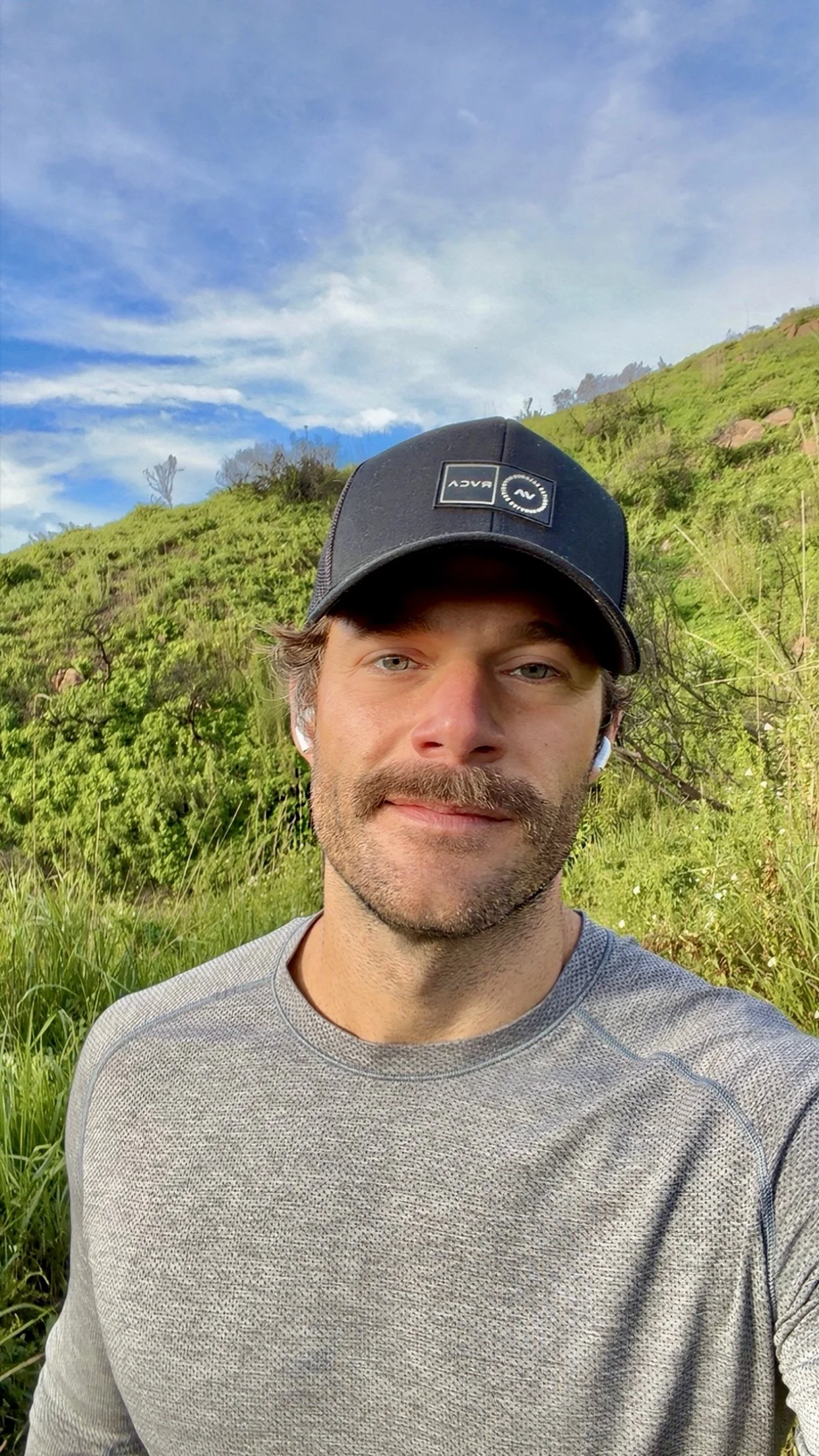 A man with a beard and mustache wearing a gray athletic shirt, a black cap, and wireless earbuds, taking a selfie outdoors with green hills and a blue sky in the background.