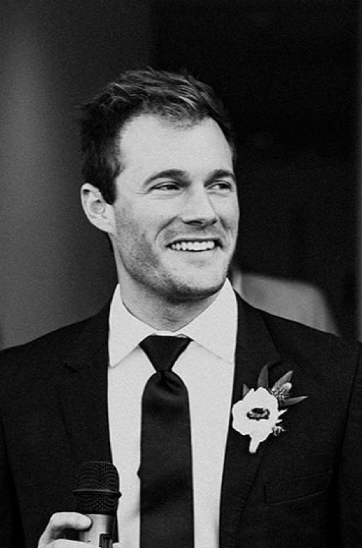 A young man in a tuxedo with a boutonniere, smiling, holding a microphone, possibly speaking at an event.