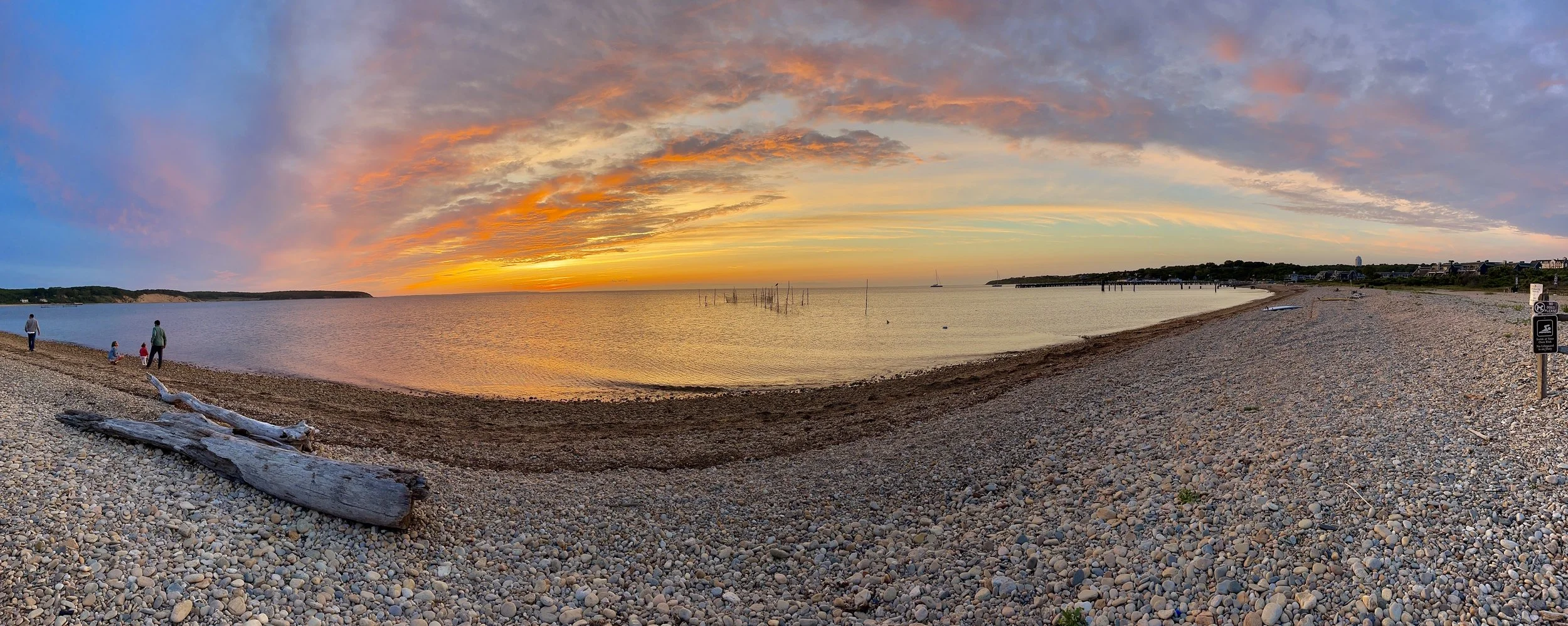 A pebble beach during sunset with a colorful sky. Few people walking, driftwood on the shore, and sailboats in the water.