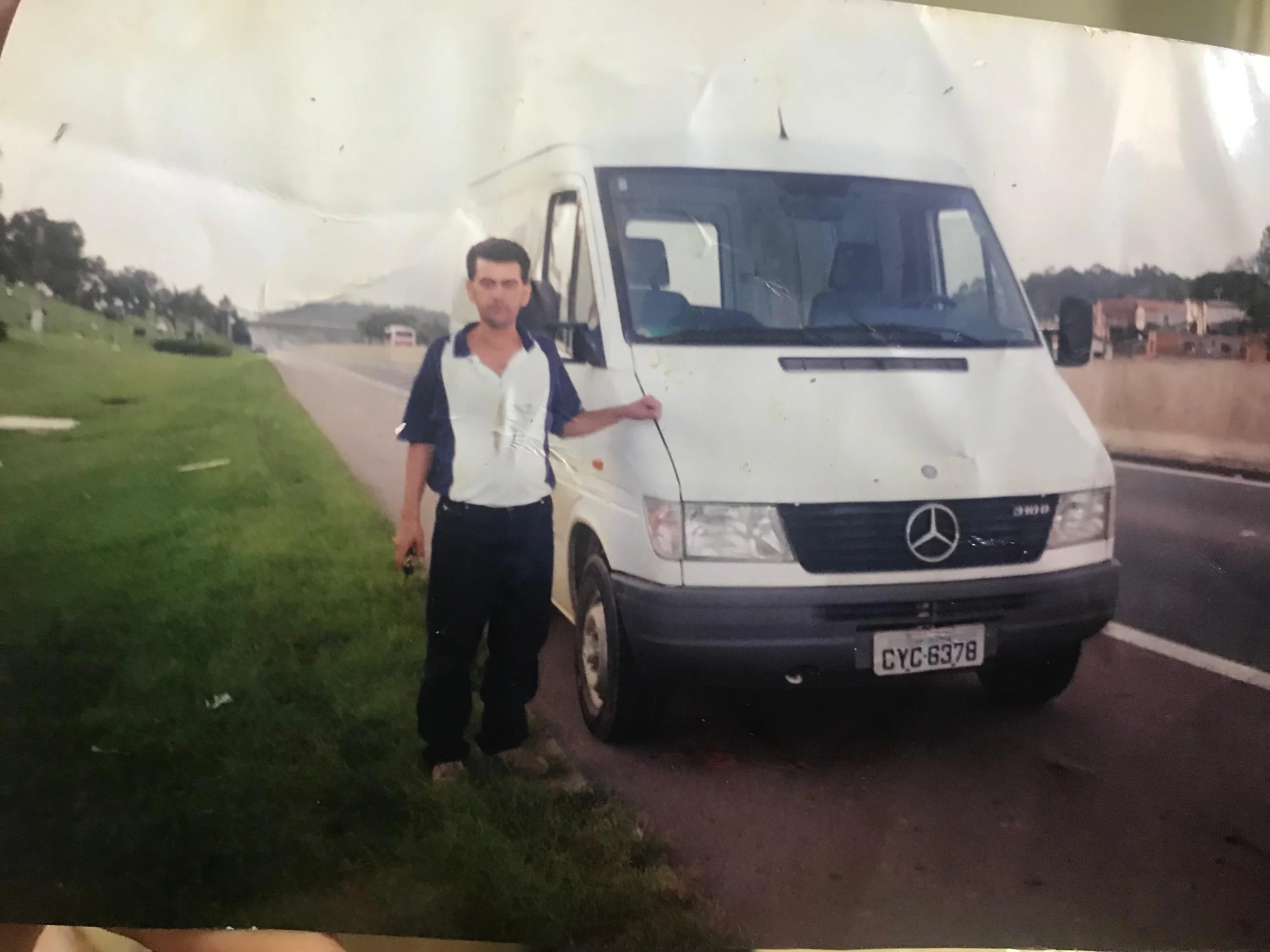 Homem de camiseta branca e jaqueta azul escura ao lado de um micro-ônibus branco na estrada, com áreas verdes ao lado e algumas construções ao fundo.
