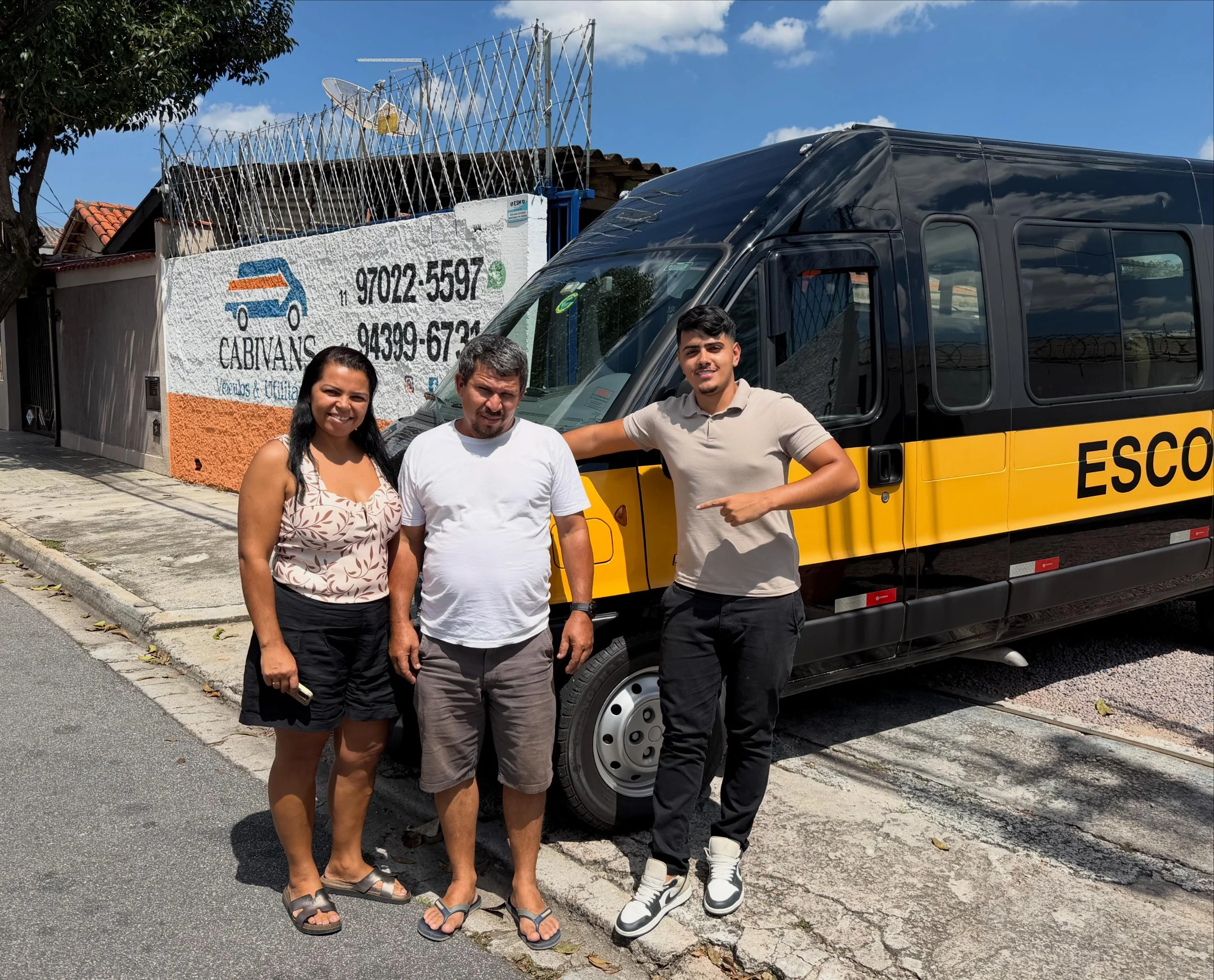 Três pessoas posando na rua em frente a uma van amarela, com uma cerca de ferro no fundo e uma parede com um cartaz de propaganda de serviços, sob um céu azul com algumas nuvens.