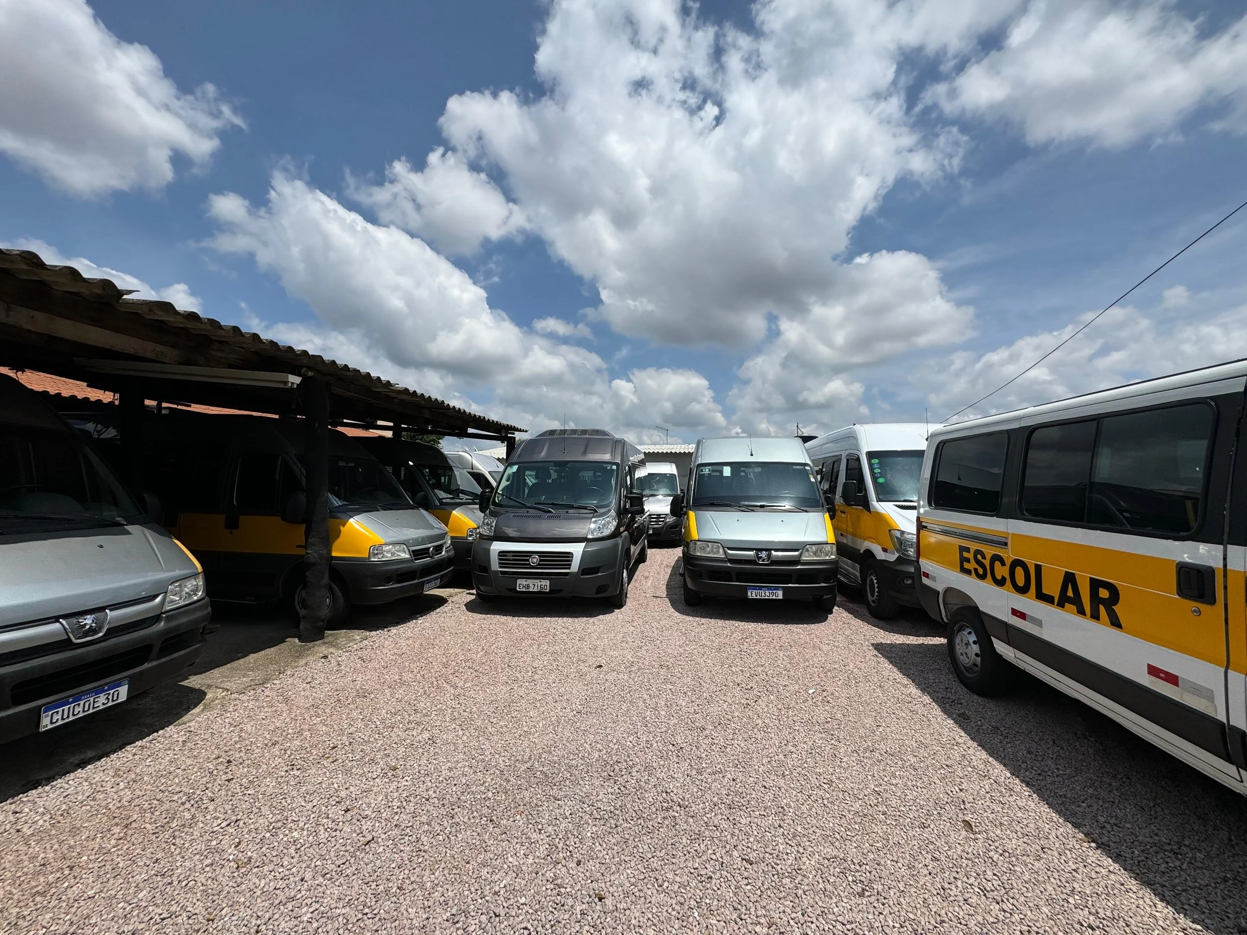 Estacionamento com vários veículos, incluindo vans escolares com a palavra 'ESCOLAR' escrita na lateral, sob céu parcialmente nublado.