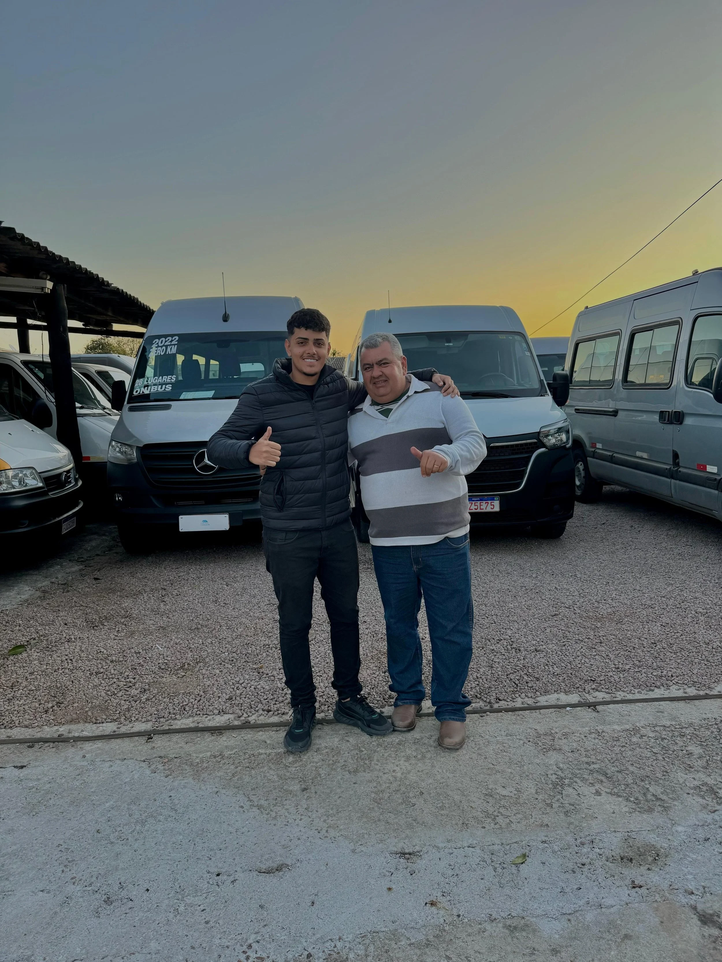 Dois homens jovens sorrindo e fazendo sinal de positivo, segurando as mãos um do outro, de frente para uma fila de veículos, ao pôr do sol, em uma área de estacionamento ao ar livre.