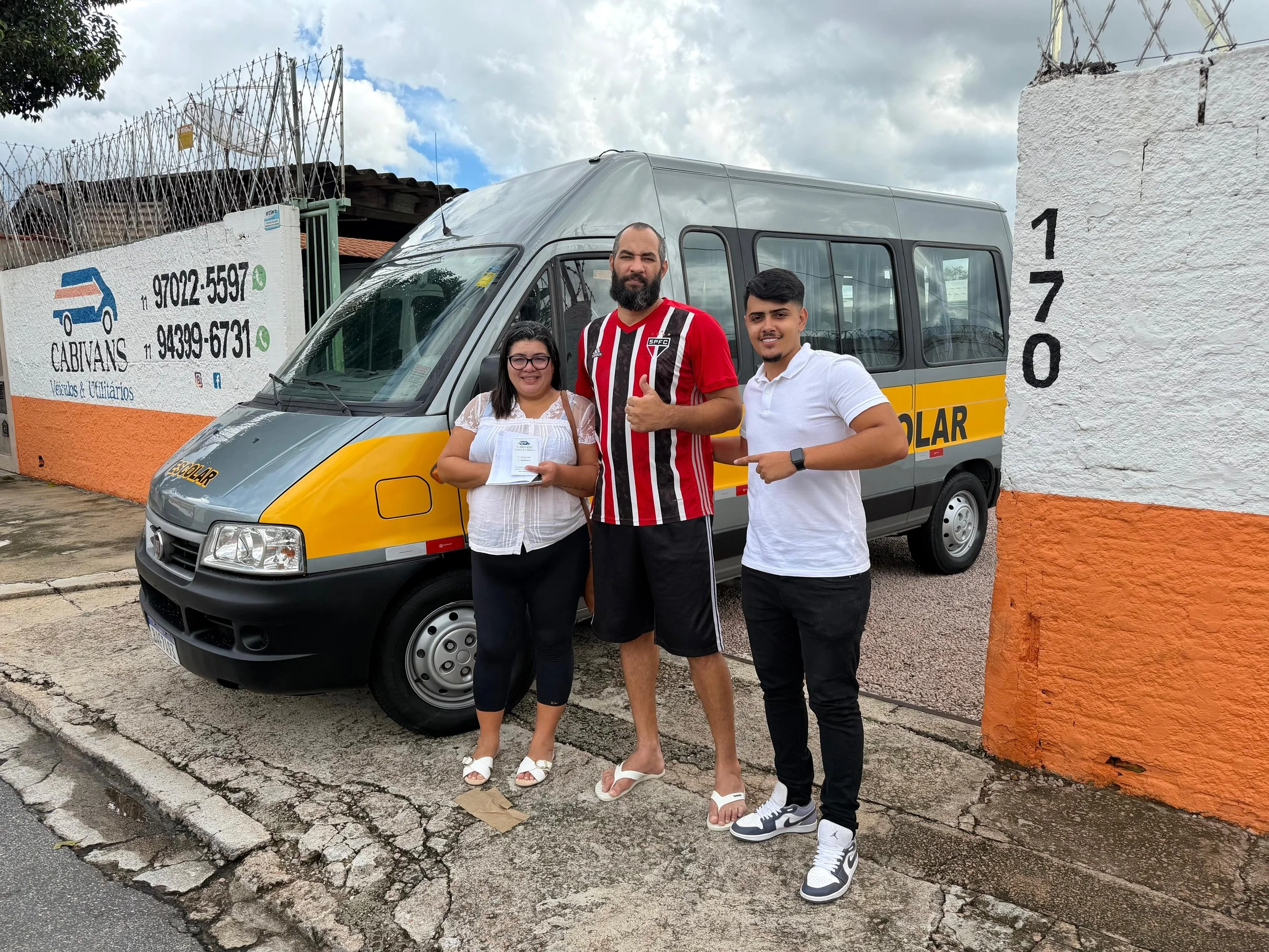 Três pessoas posando na rua na frente de um veículo de transporte escolar amarelo e cinza. Duas mulheres e um homem, ambos sorridentes, com roupas casuais e dentro de um ambiente externo sob céu parcialmente nublado.