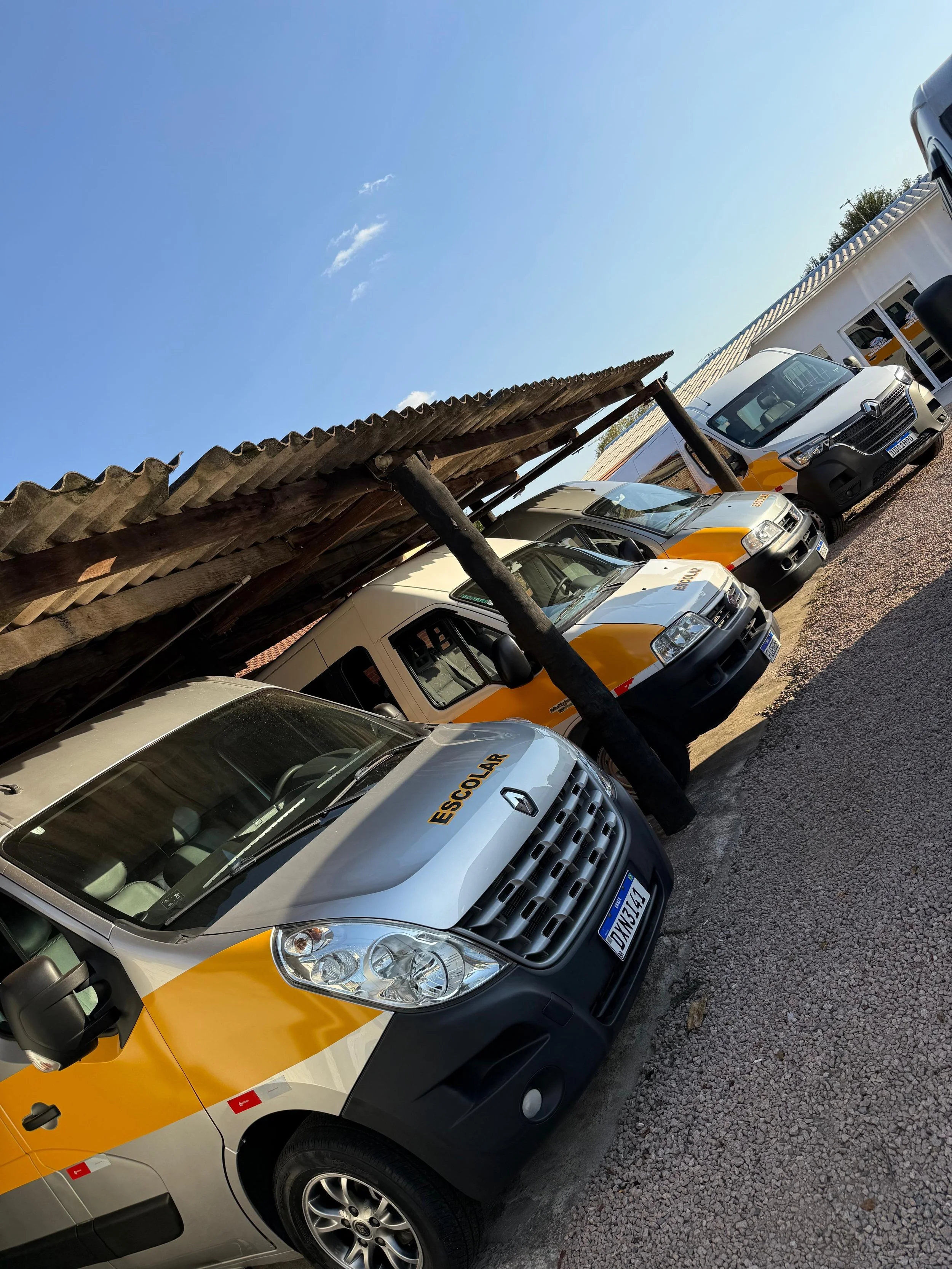 Vários veículos de transporte escolar estacionados ao ar livre, com uma estrutura de cobertura de telhado de amarrações de madeira ao fundo e céu claro.
