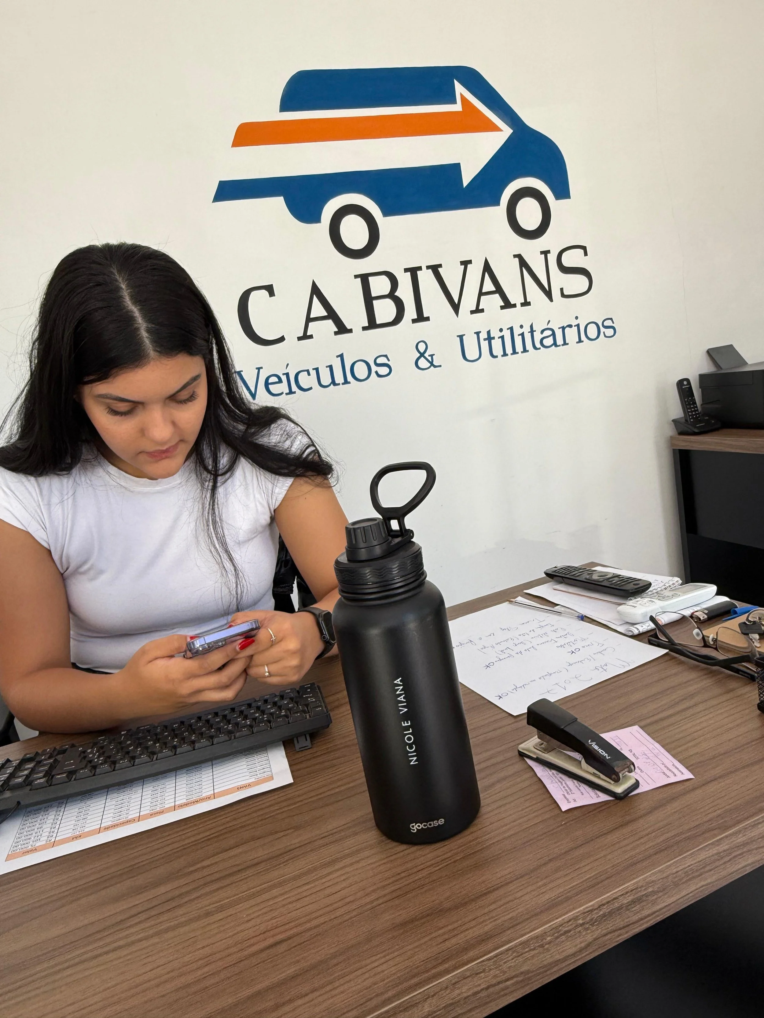 Pessoa usando celular em uma mesa de escritório, com certificados, documentos, computador, teclado, garrafa de água e telefone ao lado, com parede ao fundo com logotipo da empresa 'CAVANS Veículos & Utilitários'.