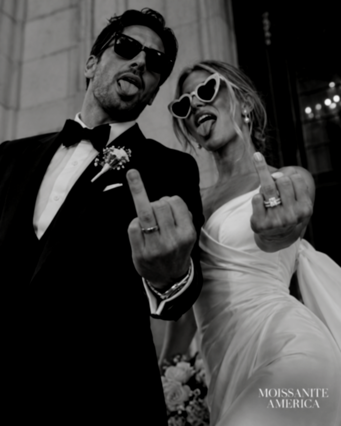 Newlyweds in sunglasses holding up their ring fingers to show a moissanite engagement ring and wedding bands