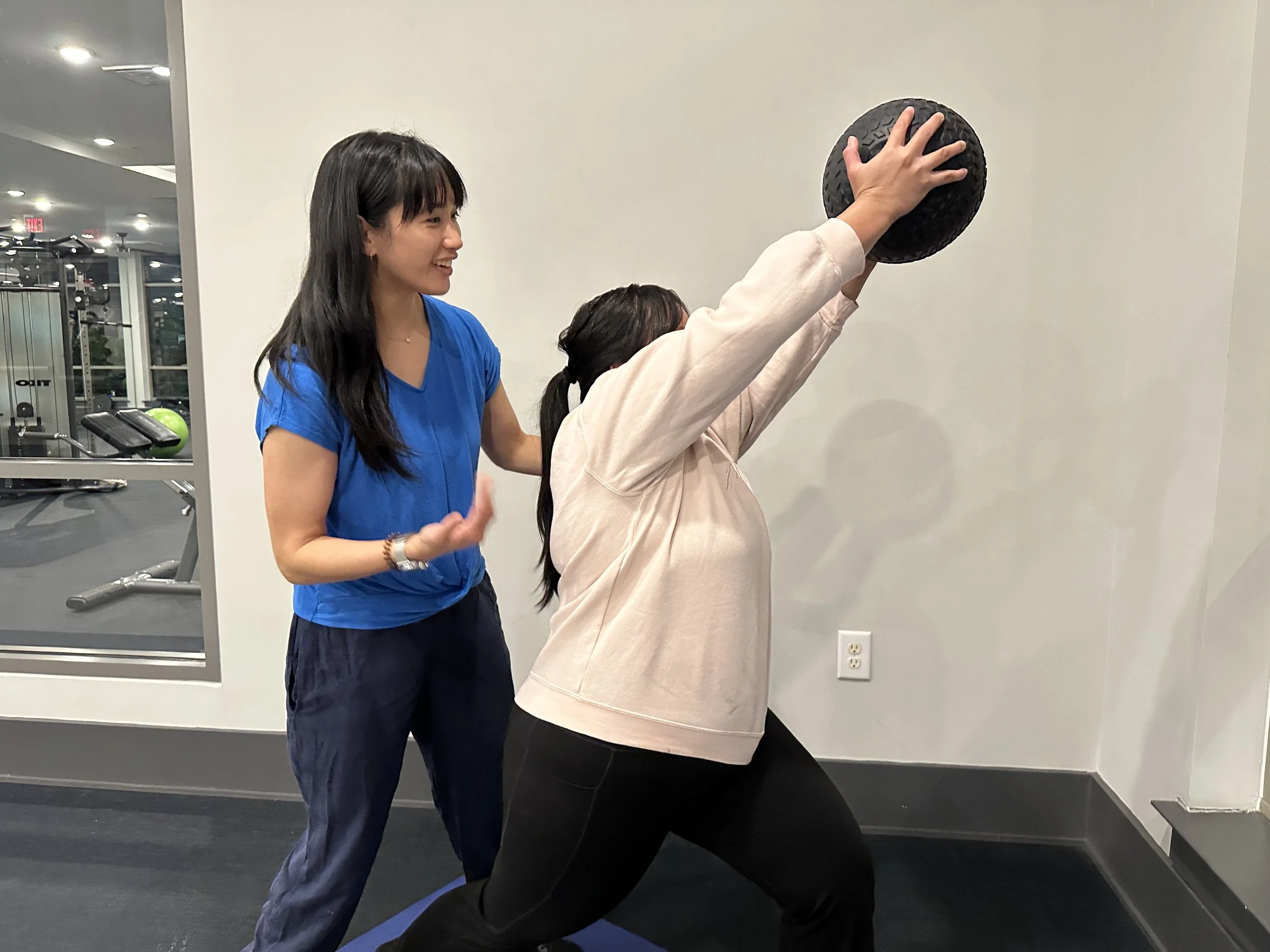 A woman in a beige hoodie is holding a black textured medicine ball above her head while another woman in a blue shirt and dark pants watches and encourages her, in a gym with mirrors and exercise equipment.