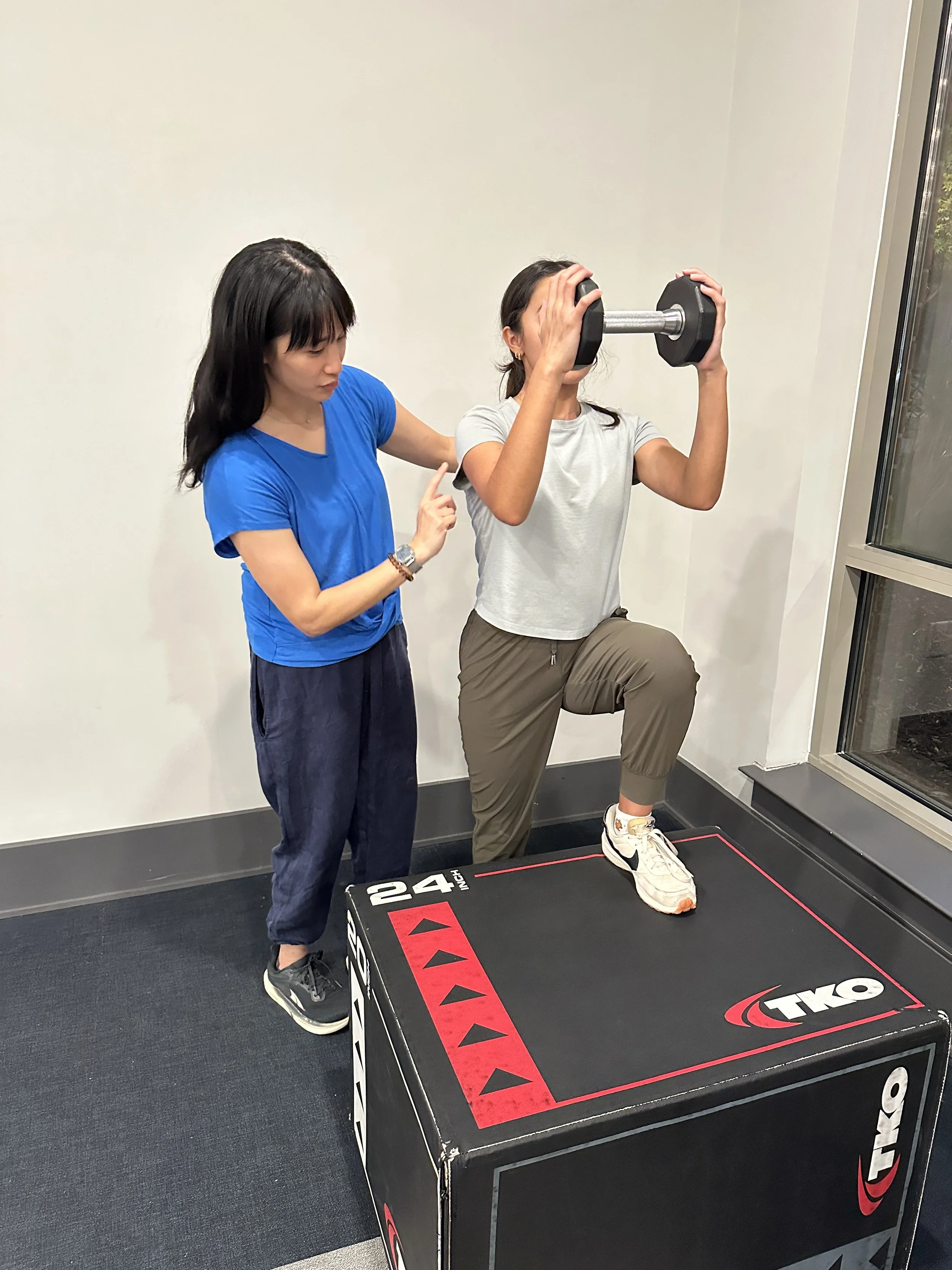 A woman in gray athletic clothes and white sneakers is doing a leg exercise by stepping onto a large black plyometric box while holding a dumbbell in front of her face. A woman in a blue shirt and dark pants is standing beside her, giving instructions or encouragement, in a brightly lit room with a plain white wall and a glass window.