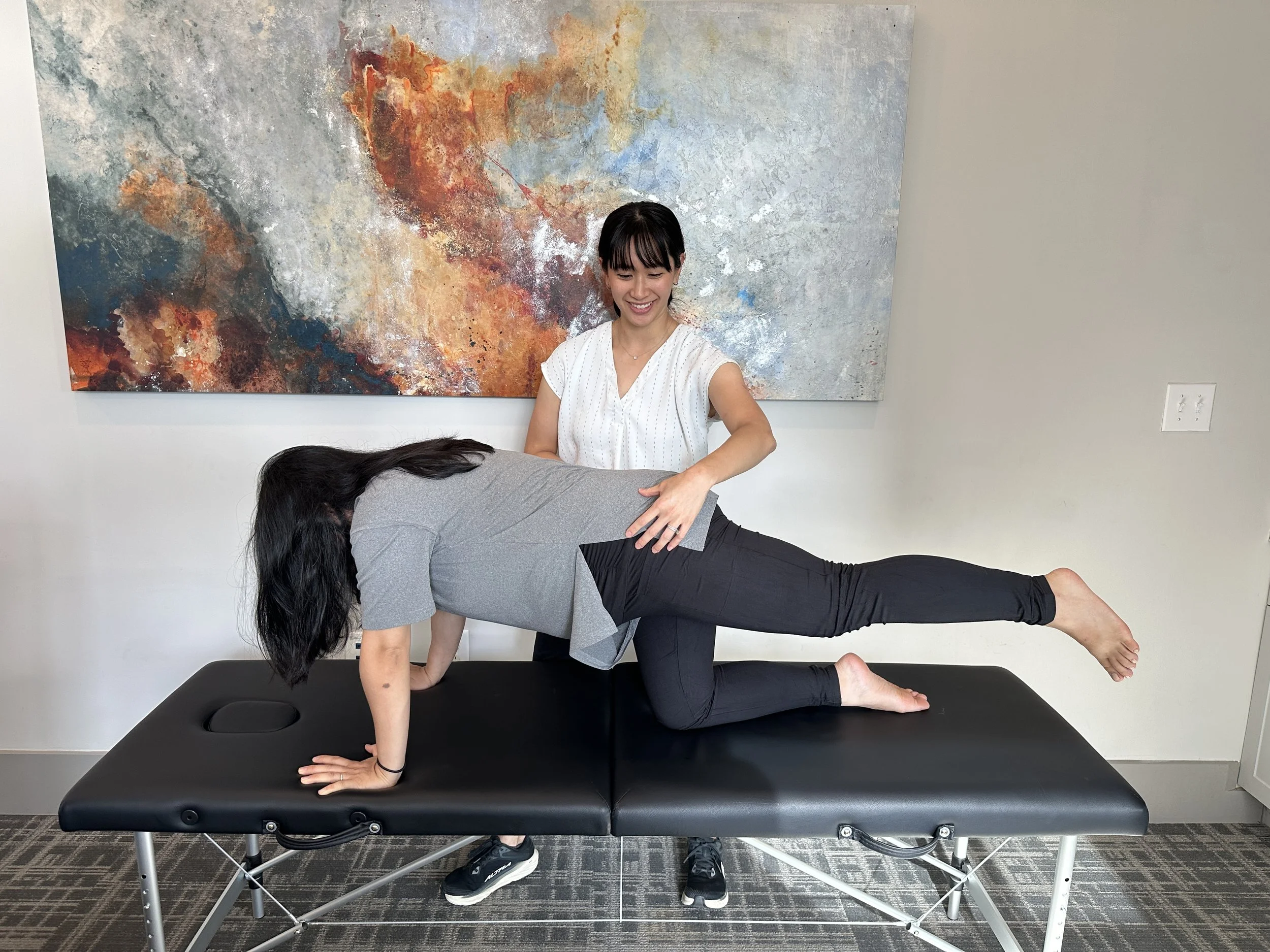 A woman in a white top practicing a yoga plank pose on a black massage table with a dark-haired woman in a gray shirt supporting her from underneath, inside a room with an abstract colorful painting on the wall.
