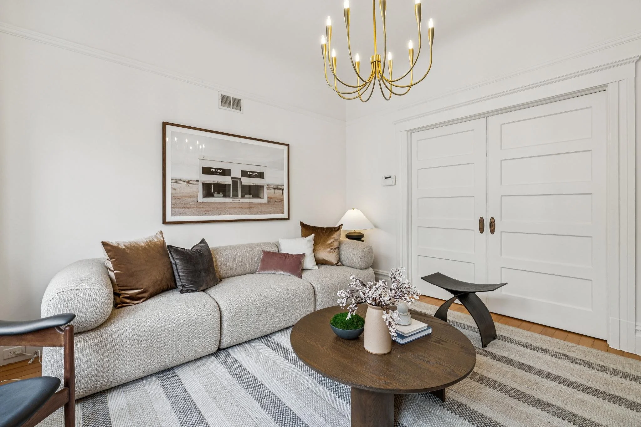 Living room with beige sofa decorated with various pillows, a round coffee table with vases and books, a black curved bench, a large wall mirror, and a white sliding door. A gold chandelier hangs from the ceiling.