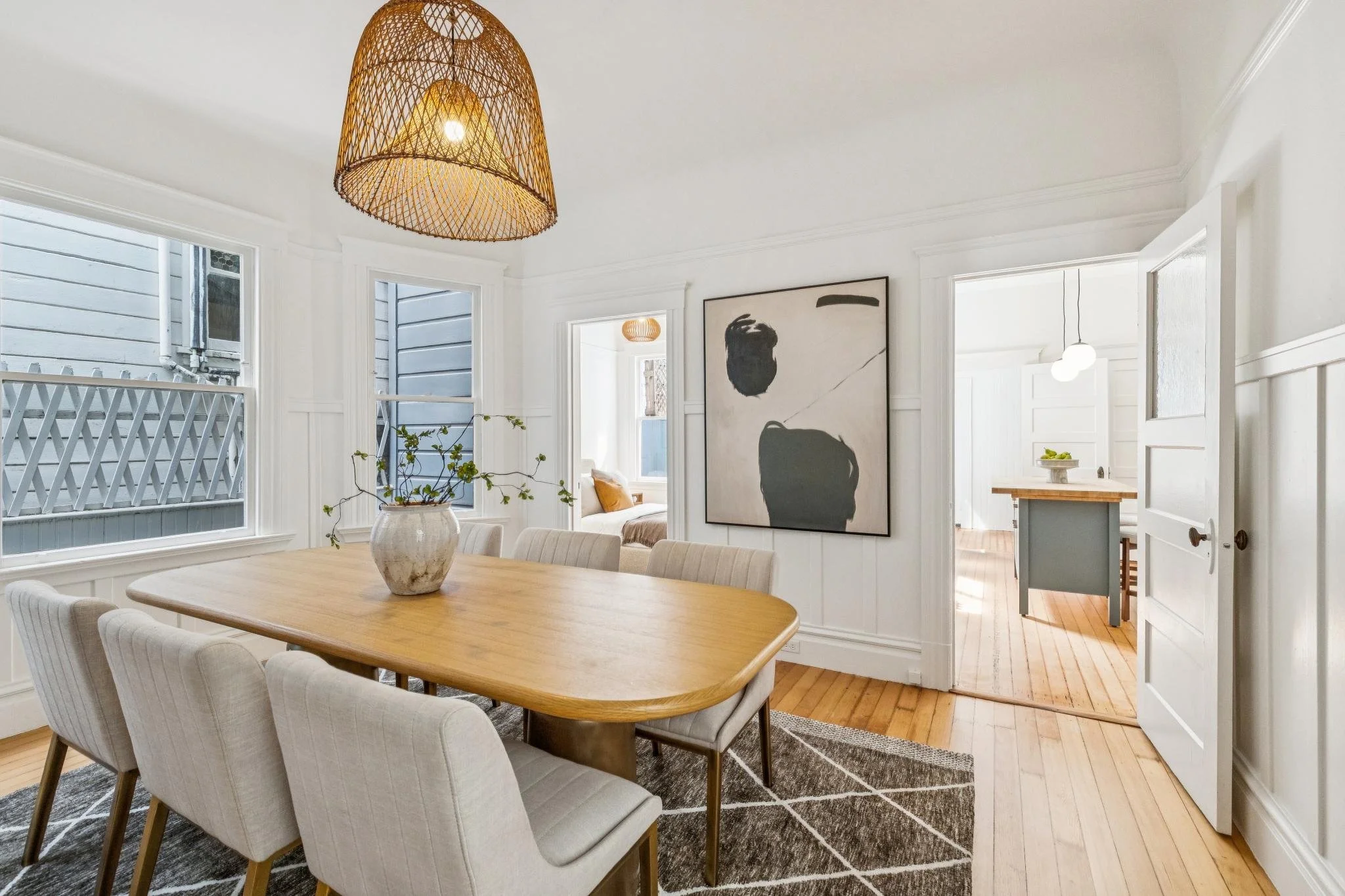 A bright, airy dining room with a wooden table and six upholstered chairs, featuring a beige and white color scheme, modern artwork, large windows with views of neighboring houses, and wicker pendant lighting.