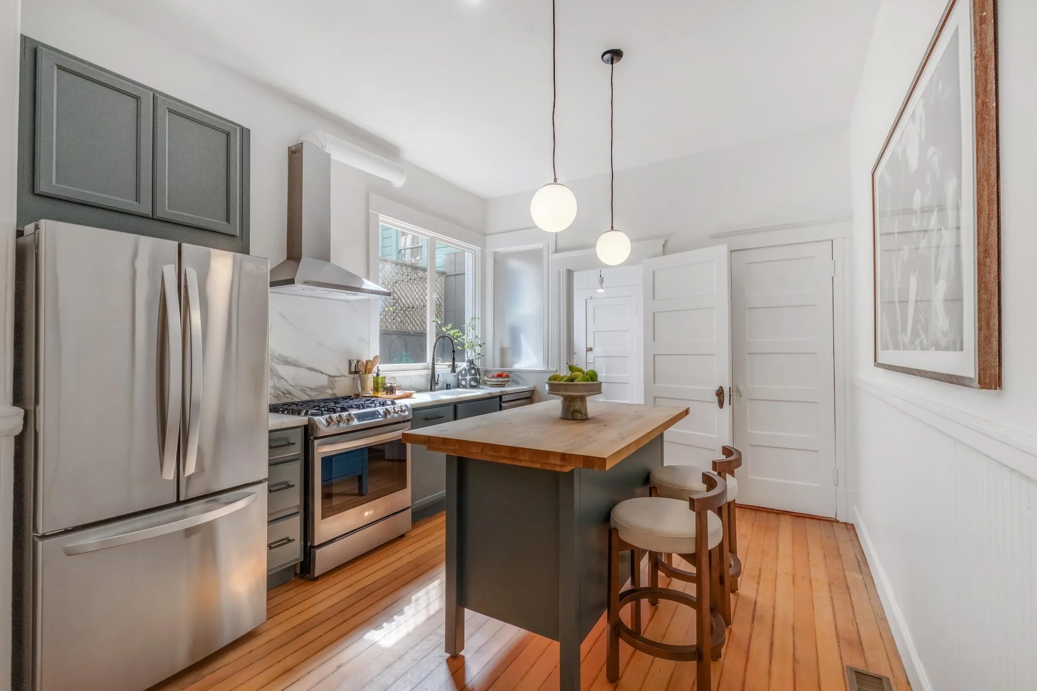 A kitchen with a white refrigerator, a stove with a marble backsplash, pendant lights hanging from the ceiling, a window behind the sink, white paneled walls, and a wooden floor. There is a small island with chairs and a bowl of green apples on top.