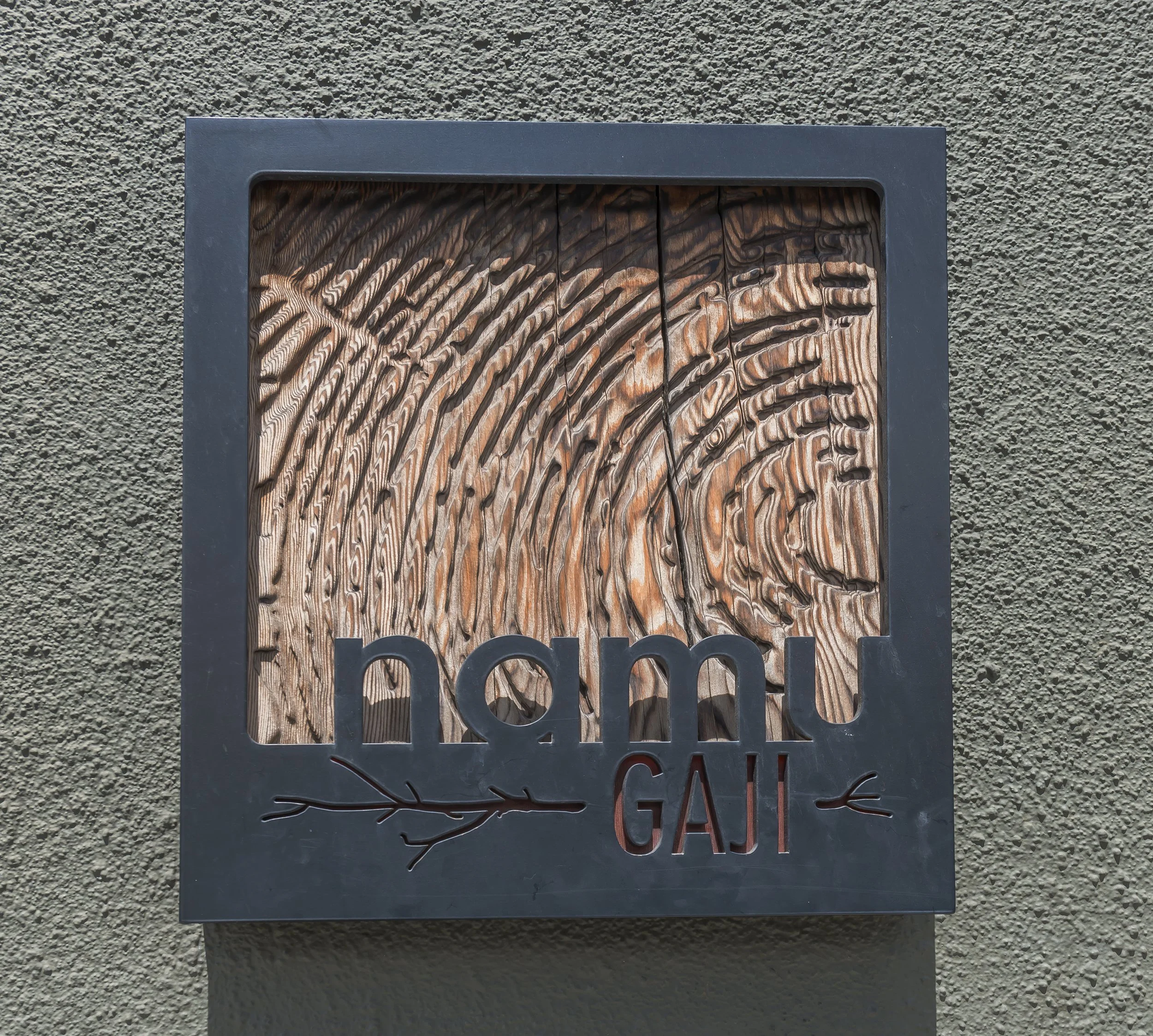 Metal sign with a tree design and partial text, mounted on a textured outdoor wall.