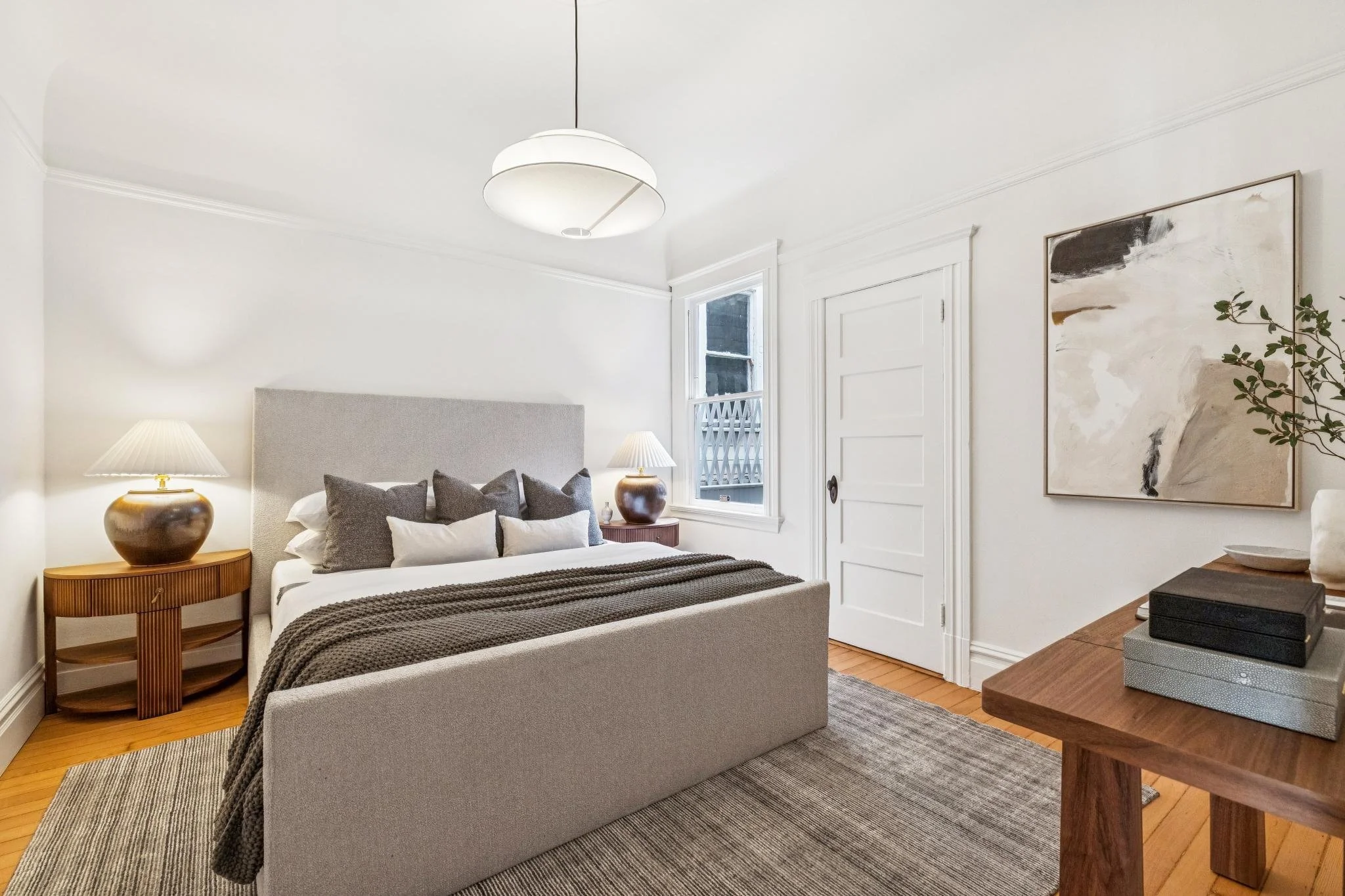 A bedroom with a bed, bedside tables with lamps, window, painting, and wooden furniture.