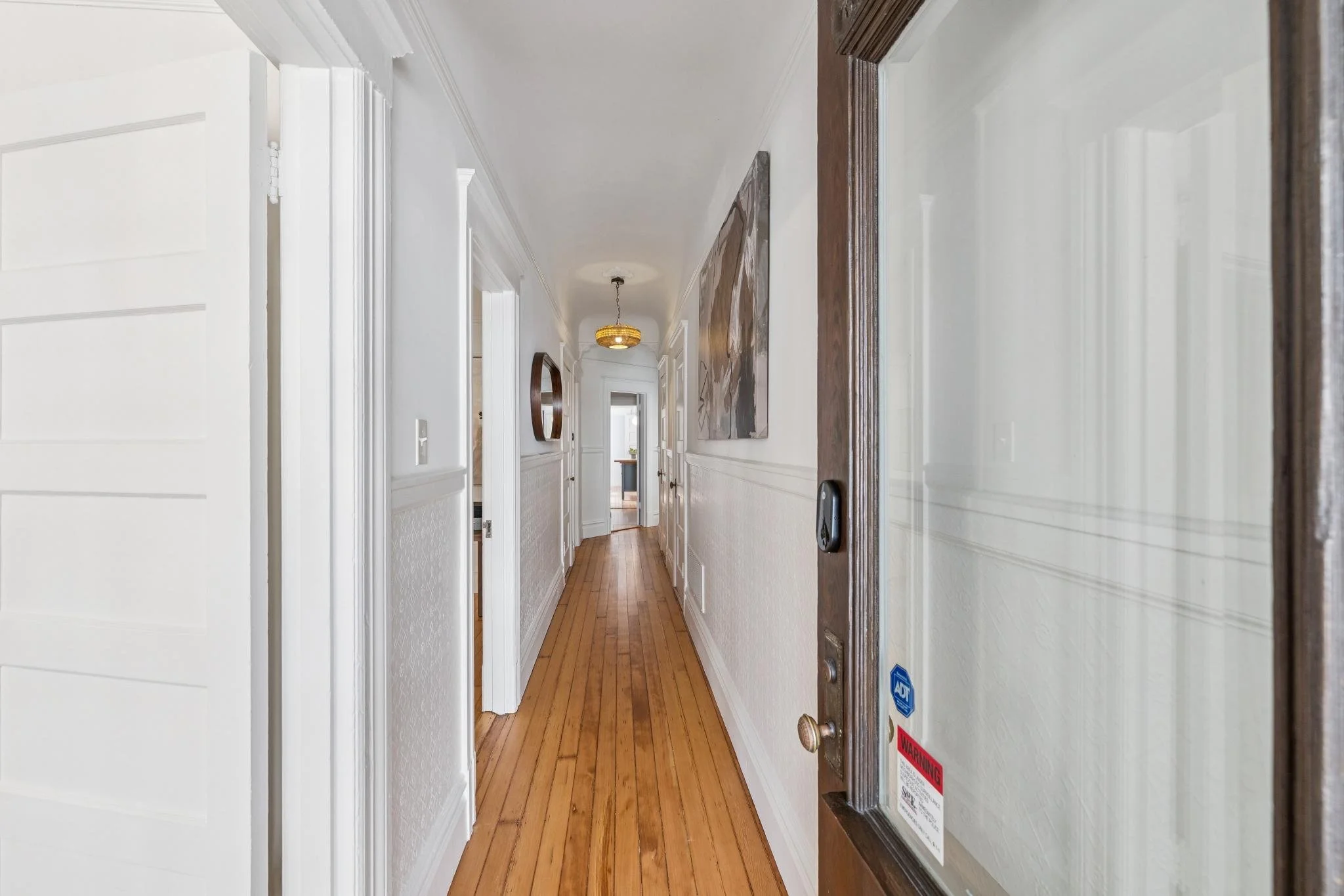 A hallway with wooden floors, white walls, and doors on both sides, with a round chandelier hanging from the ceiling at the end.