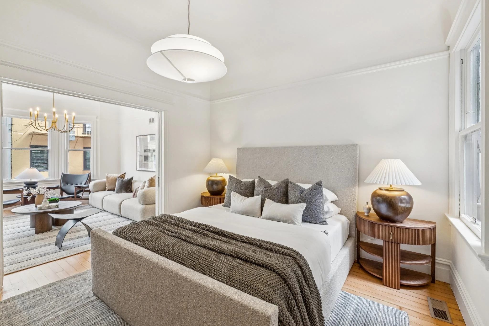 A cozy living room with large windows, a neutral color palette, and a bedroom with a bed and side tables with lamps.