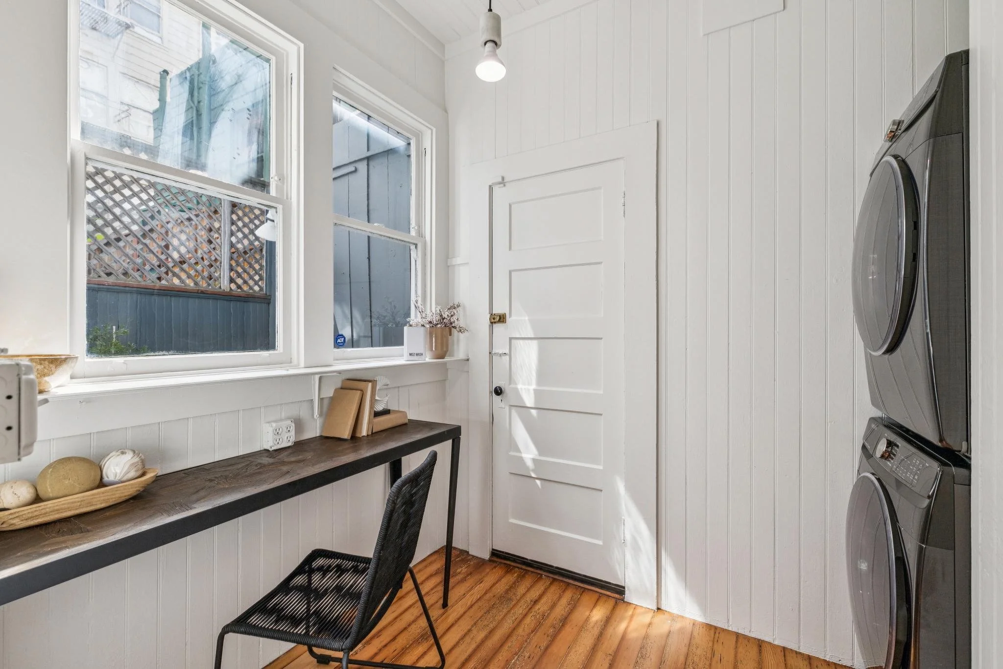 A laundry room with white wood-paneled walls, two large windows, a dark wood countertop, a black chair, and a stacked washer and dryer in a modern farmhouse style.