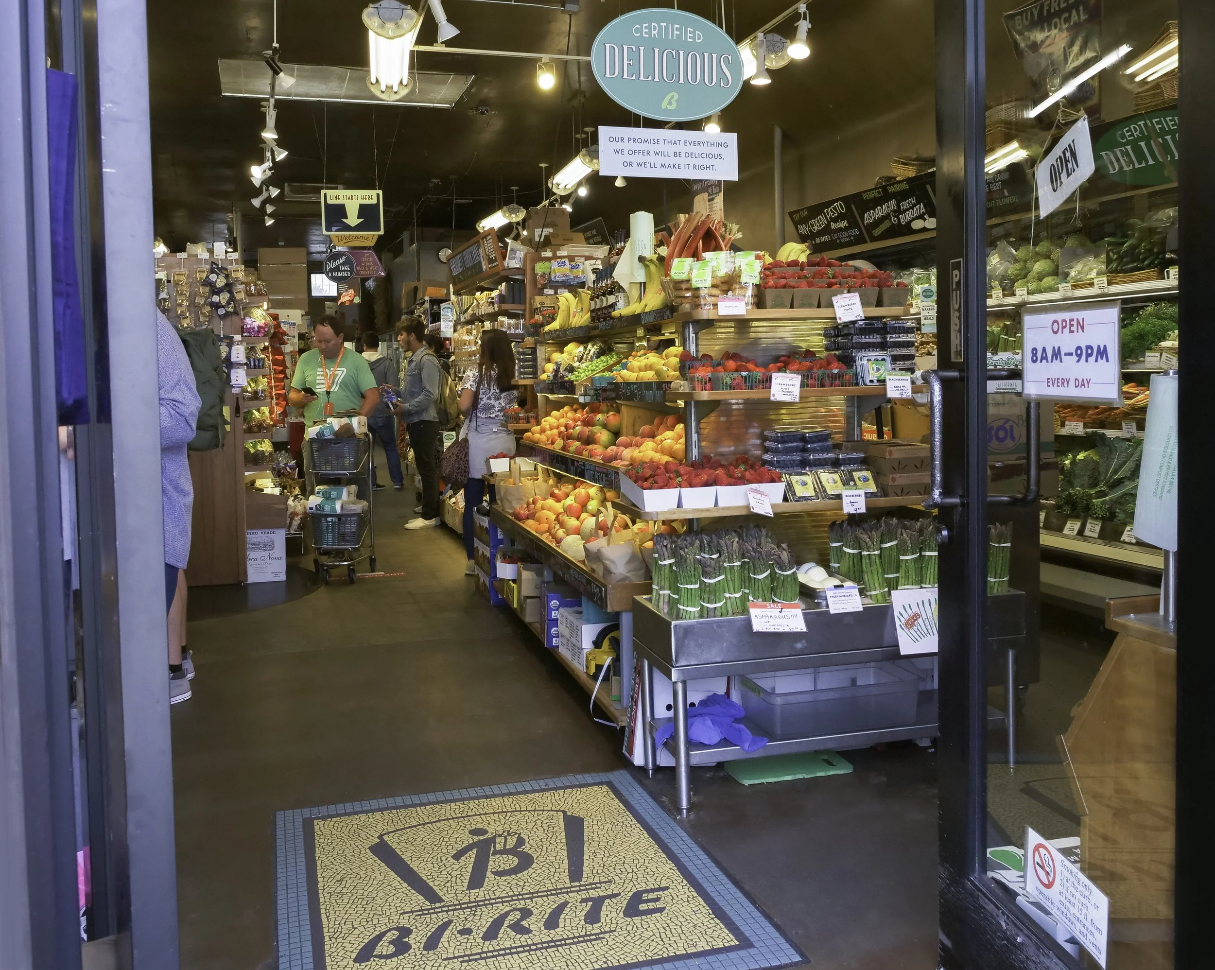 View inside a grocery store with fresh produce including strawberries, bananas, apples, and asparagus. Signs indicate store hours from 8 AM to 9 PM daily, and a large sign reads 'Certified Delicious'. The floor has a mosaic with a Bitcoin symbol. Cus