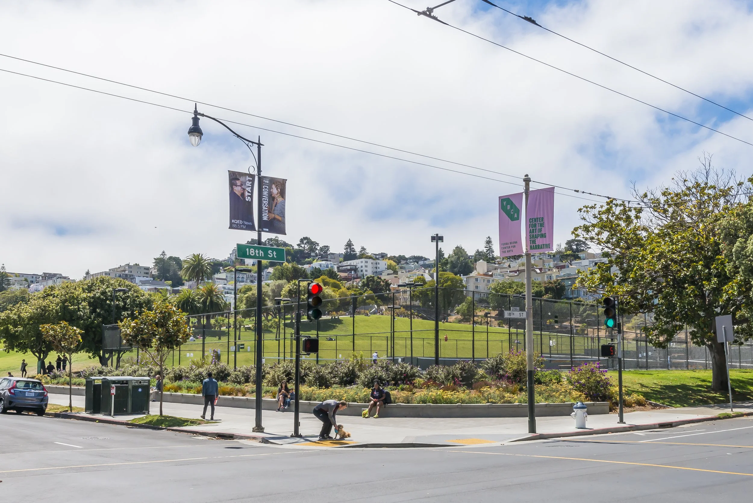 A city park with a grassy field, walking paths, and a tennis court. People are walking and sitting in the park. There are trees, bushes, and flowers surrounding the area. Streetlights, traffic signals, and banners hang above the streets. Hills with h