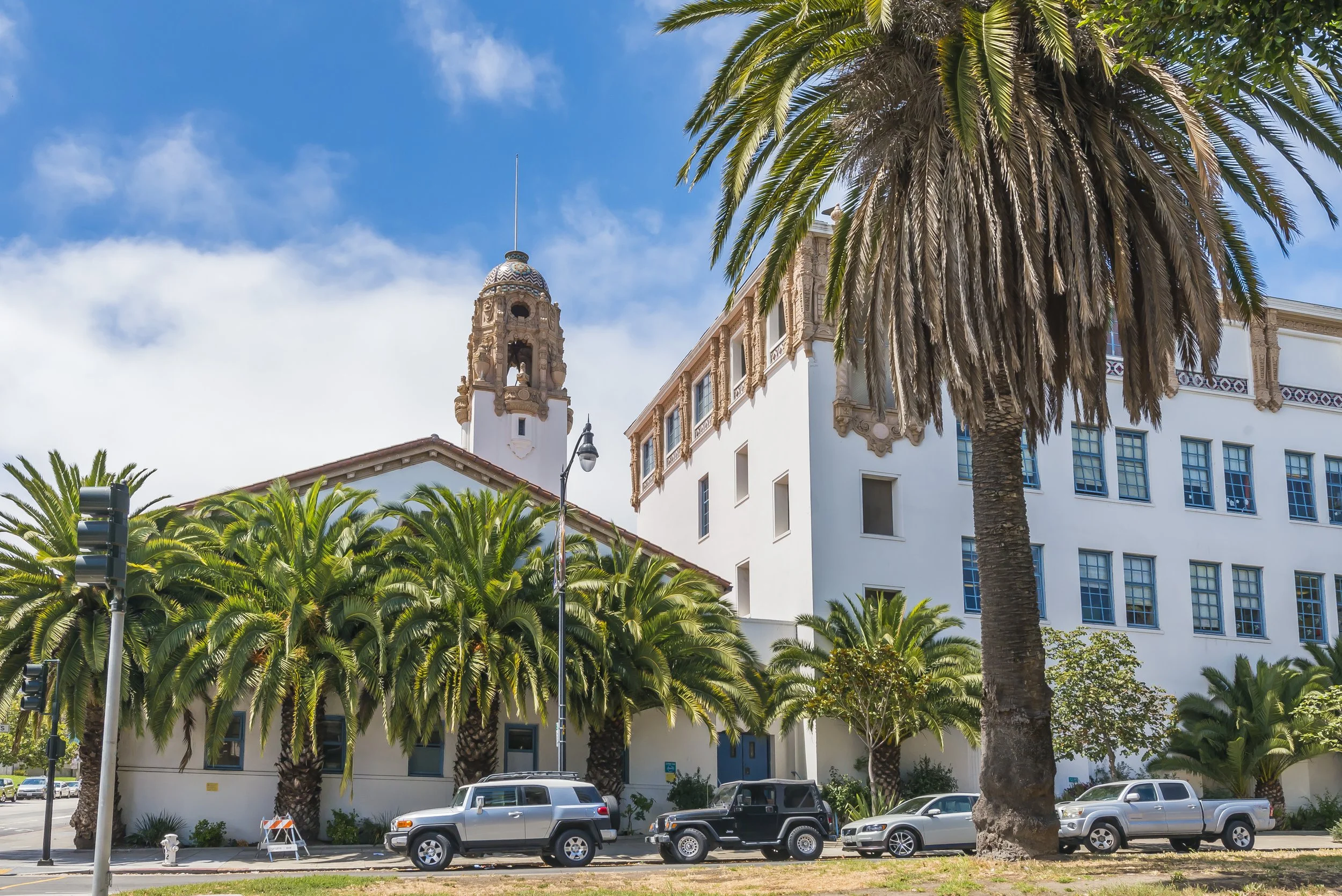 A street scene featuring a large palm tree, a white building with decorative architectural details, a clock tower, and parked cars under a partly cloudy blue sky.