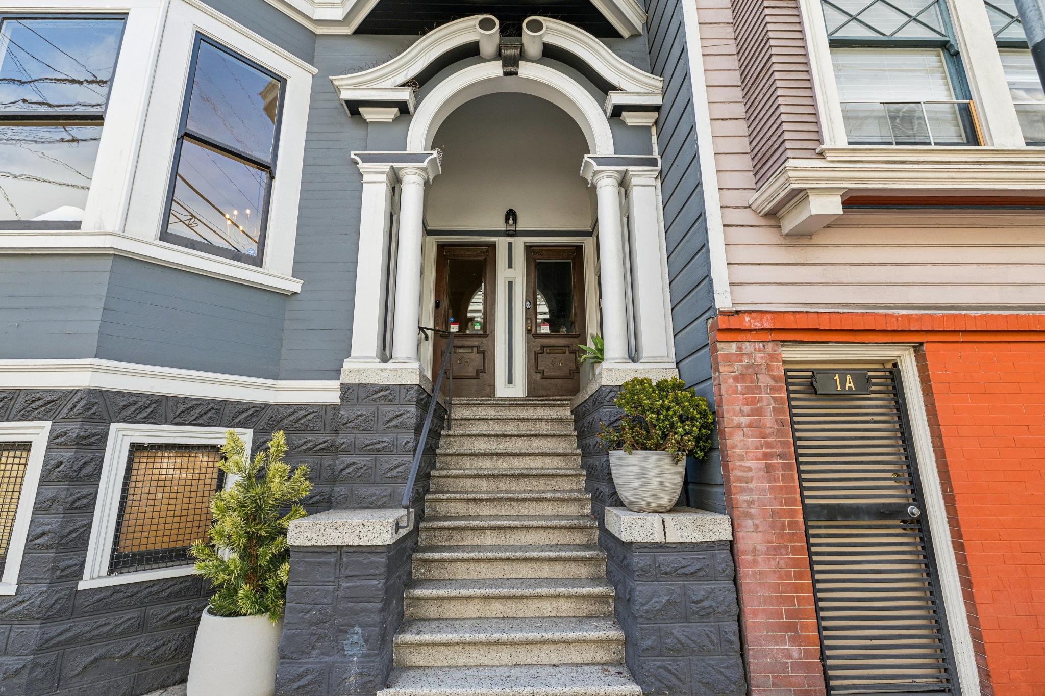 Front entrance of a modern two-story house with an arched doorway, stone stairs, and decorative columns. The house features dark gray and brick exterior walls, large windows, and potted plants flanking the stairs.