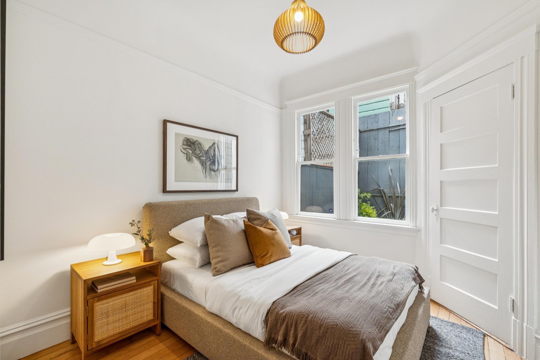 A bedroom with a bed, brown and white bedding, pillows, a wooden bedside table, a small white lamp, a framed black and white sketch on the wall, large windows showing outdoor greenery, and a white door.