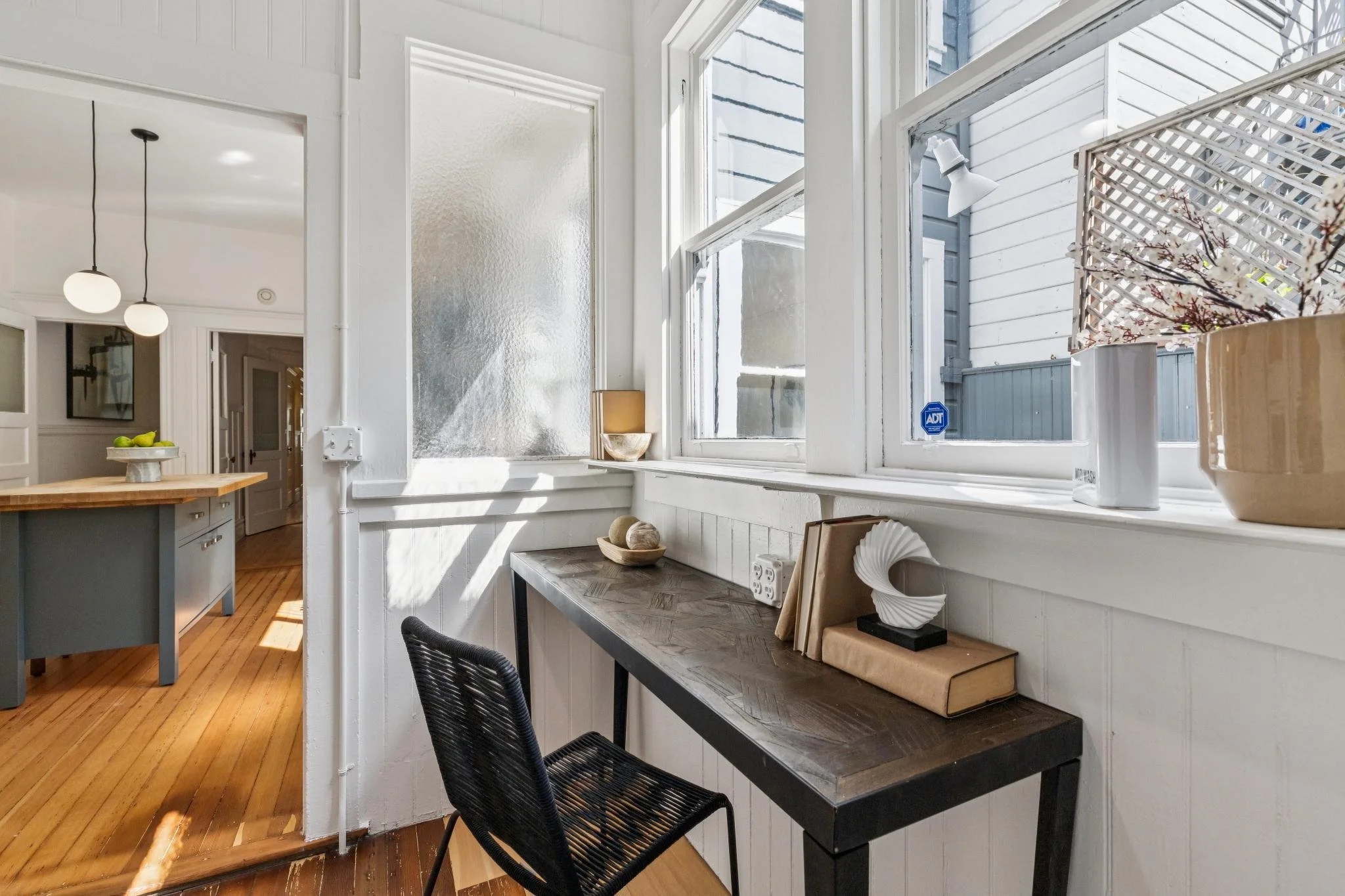 A sunlit corner of a kitchen or dining area with white walls and windows, featuring a black desk with decorative items, and a view of a neighboring house's exterior with white siding, a lattice fence, and flowering branches.
