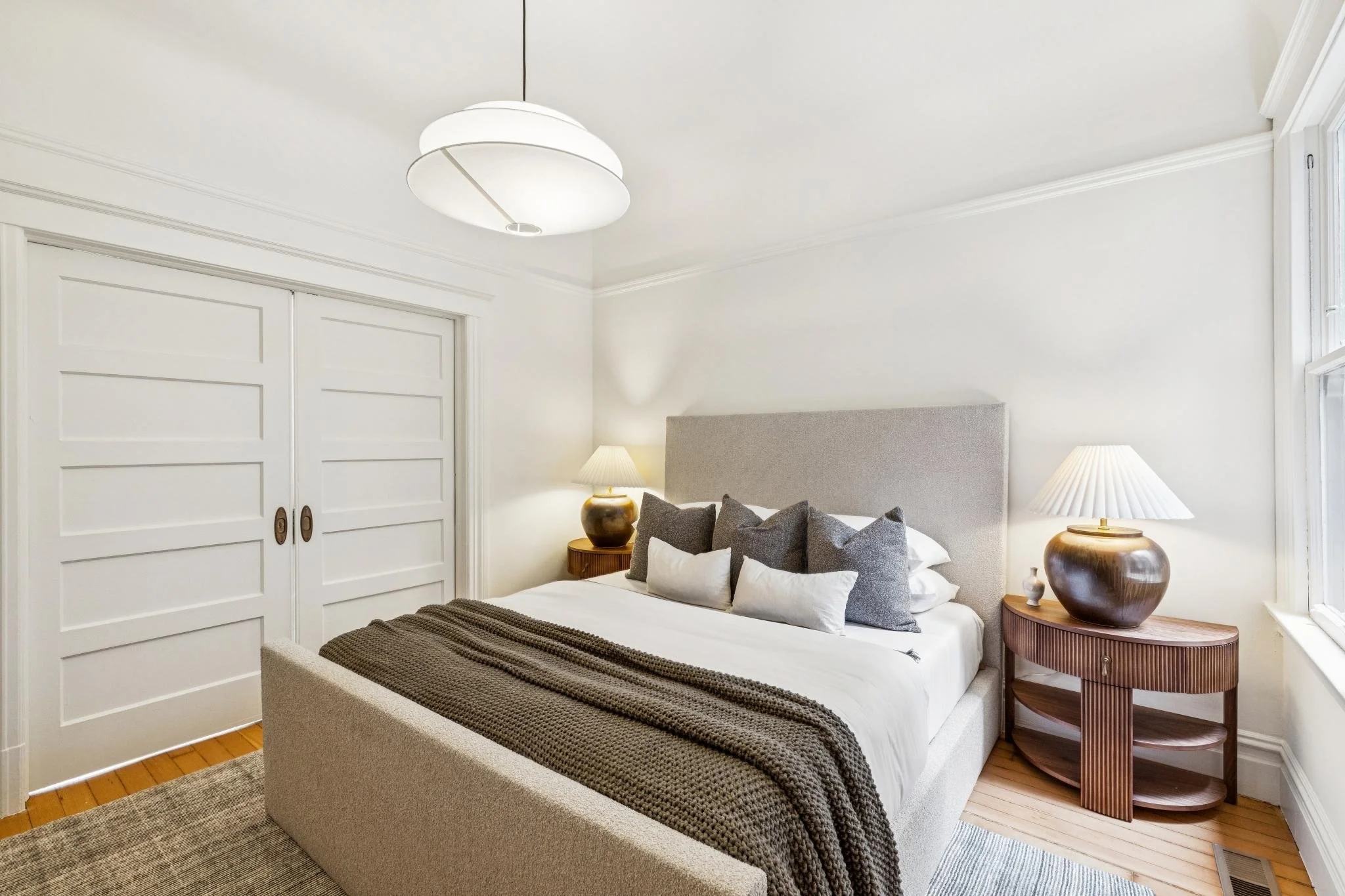 A cozy bedroom with a bed, gray upholstered headboard, multiple pillows, and a gray blanket. On either side of the bed are wooden nightstands with ceramic lamps, and a large white ceiling light hangs from the ceiling. The room has white walls, white 