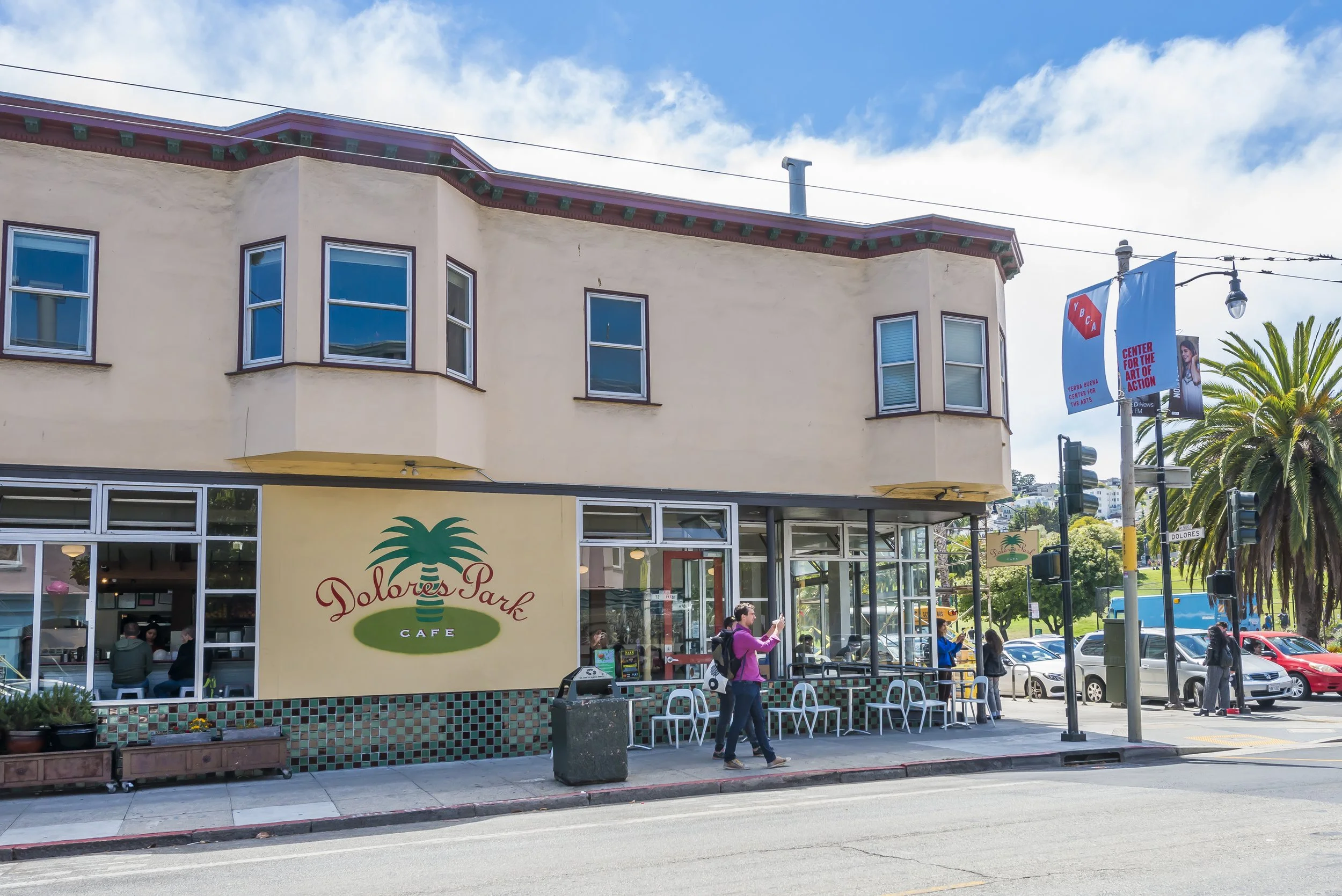 Street view of Dolores Park Cafe with people sitting outside, people walking by, and a yellow building with a sign for the cafe featuring a palm tree logo. There are palm trees, a light pole with banners, and parked cars along the sidewalk under a pa