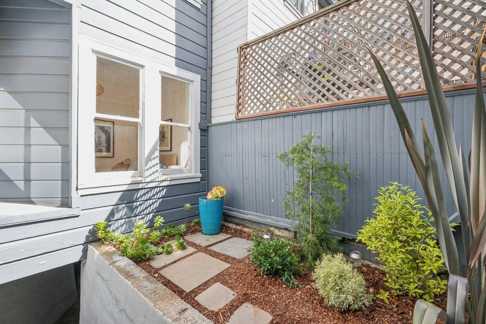 View of a small backyard garden with plants, a blue pot with succulents, stone pathway, and a wooden privacy fence next to a house with blue siding and a window.