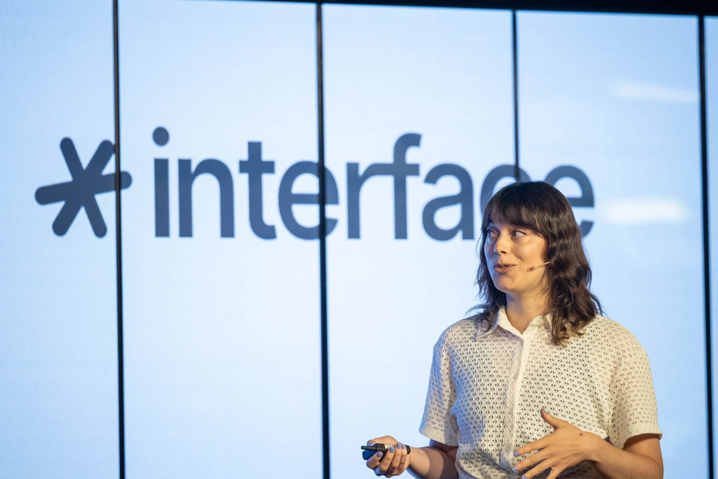 Une femme debout devant un grand écran affichant le logo de Facebook. Elle tient un téléphone portable dans la main droite et semble en pleine présentation ou en discours.