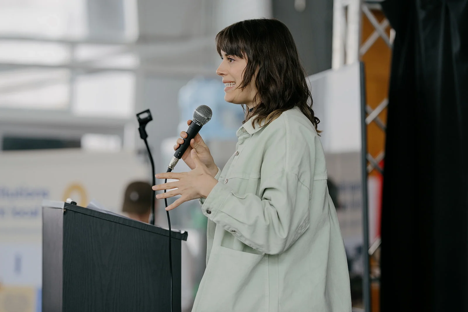 Femme parlant lors d'un discours avec un microphone devant un pupitre, dans un environnement intérieur, probablement un centre de conférence ou une manifestation publique.