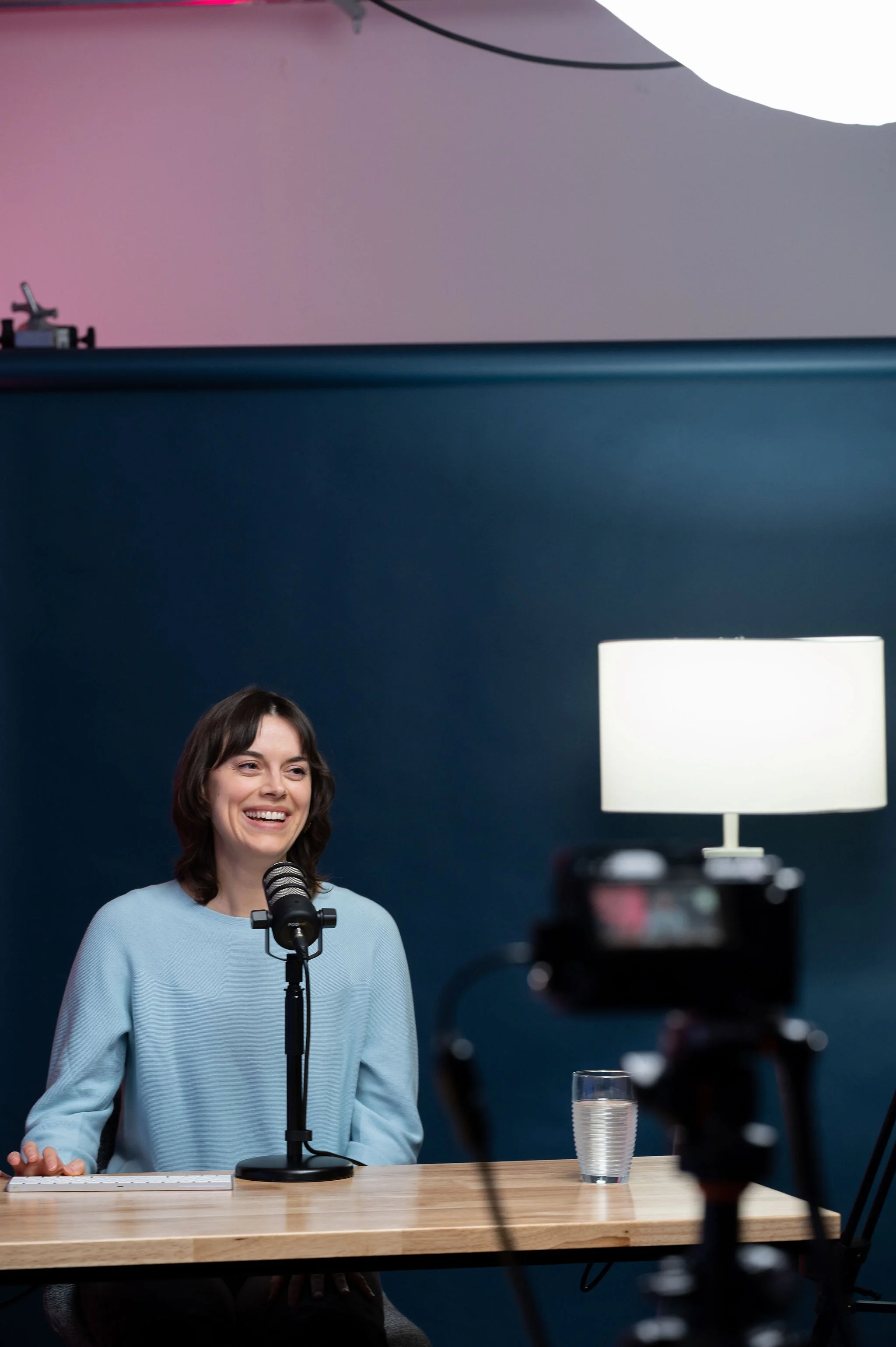 Une femme souriante, avec des cheveux bruns et courts, parle dans un microphone lors d'une interview ou un podcast, assise devant un ordinateur sur une table en bois, une verre d'eau à côté, dans une pièce avec un mur bleu foncé, une lampe blanche et une caméra en première plan.