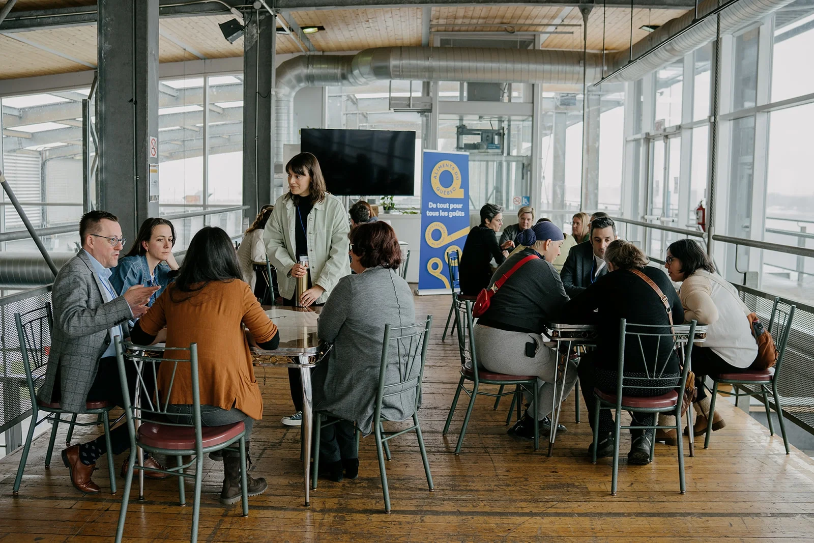Groupe de personnes assises autour de tables dans un intérieur lumineux avec vue panoramique, certaines parlant, d'autres utilisant des appareils électroniques, dans un espace moderne avec grandes fenêtres et ventilation visible.