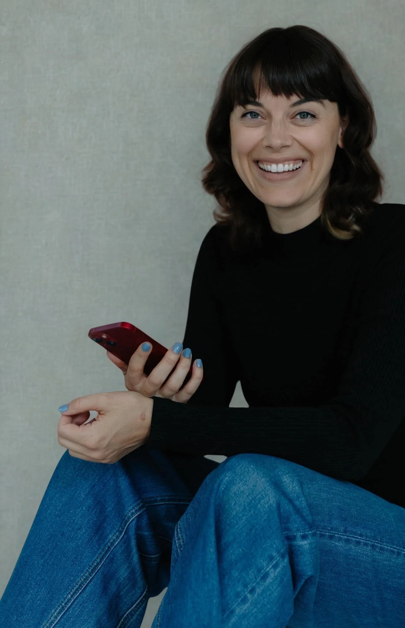 Une femme souriante assise avec des jeans et un pull noir, tenant un téléphone rouge dans sa main, dans un intérieur avec un mur beige