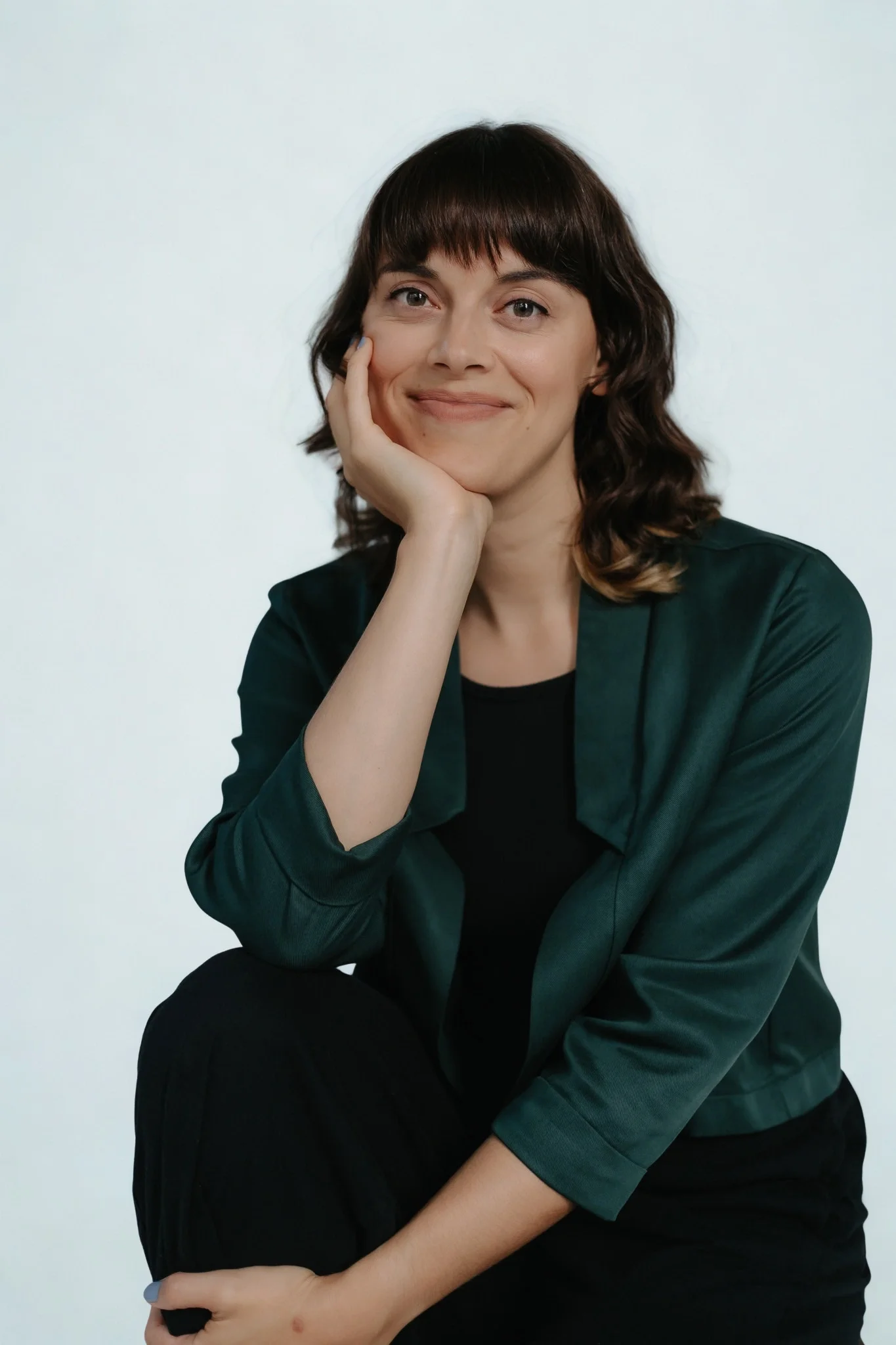 Une femme aux cheveux bruns bouclés et à la peau claire, souriante, qui repose sa tête sur sa main, portant une chemise vert foncé et un pantalon noir, assise contre un fond blanc.
