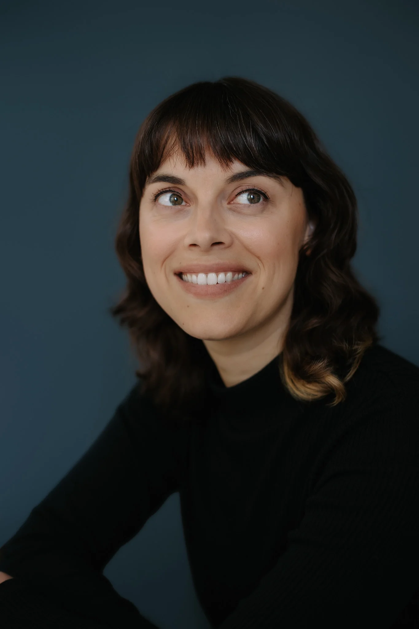 Une femme avec des cheveux bruns, souriante, portant un pull noir, posant contre un fond neutre.
