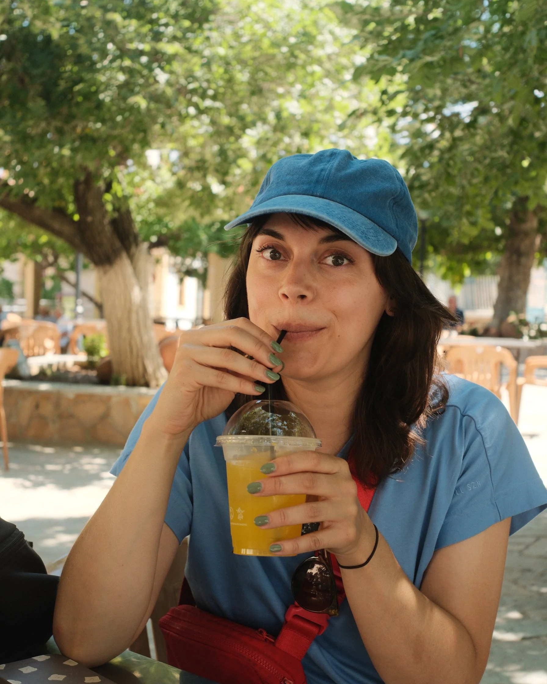 Une jeune femme avec un chapeau bleu buvant une boisson orange en plein air, sous un arbre ensoleillé.