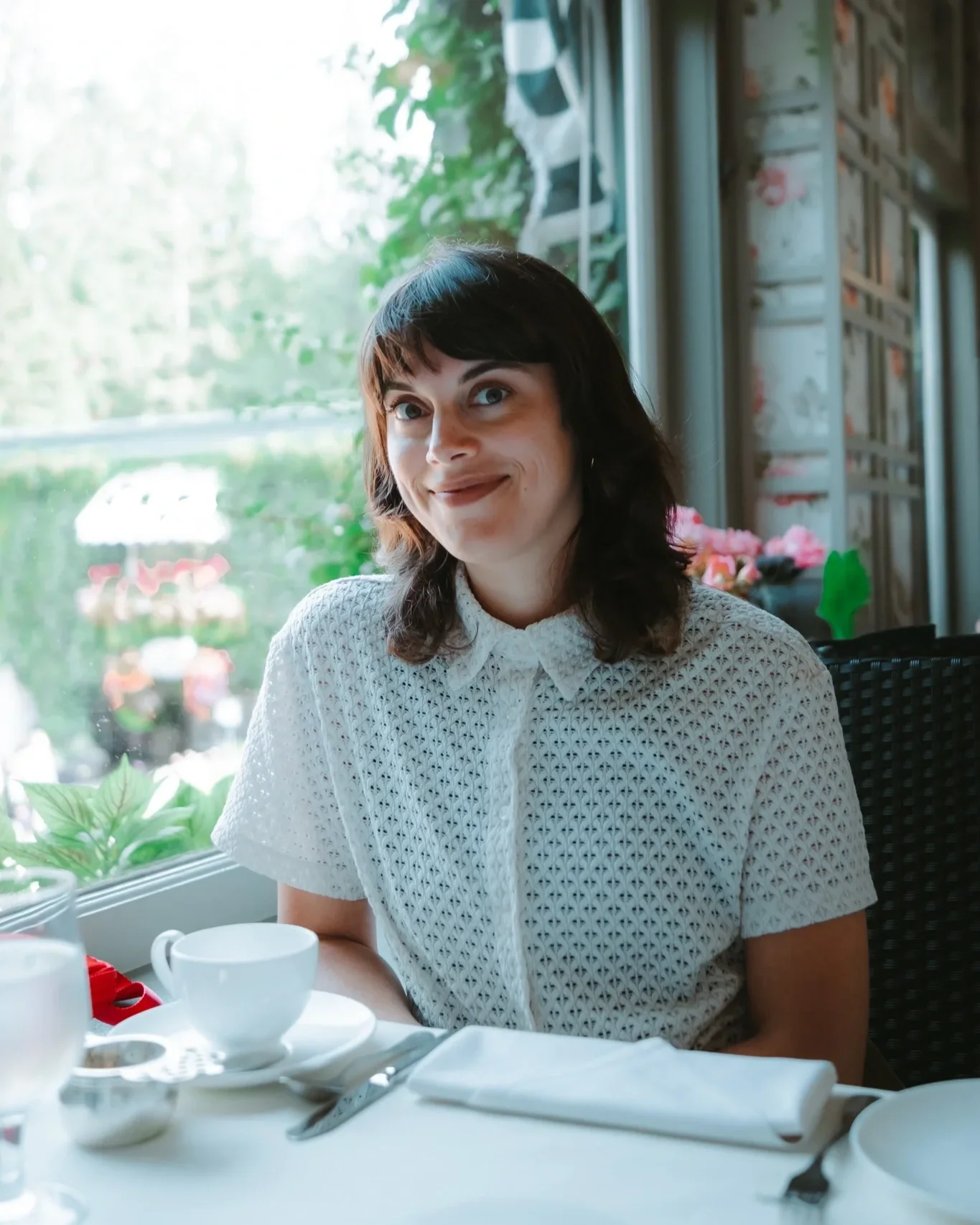 Une femme souriante, assise à une table dans un restaurant, devant une grande fenêtre avec un paysage vert extérieur, vêtements blancs à motifs, avec des tasses et assiettes sur la table.
