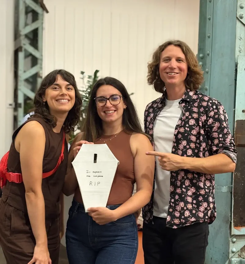 Trois jeunes adultes souriants posent ensemble, la femme au centre tient une maquette de cercueil avec l'inscription "Ici reposent mes compétences RIP". La personne à droite pointe le cercueil, le fond montre un mur et une structure métallique.