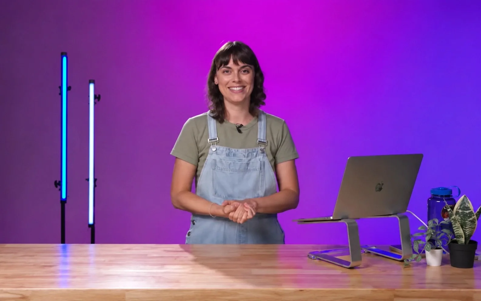 Femme souriante avec une tablette sur un bureau en bois, fond violet, plantes en pot, lampes LED, ordinateur portable.