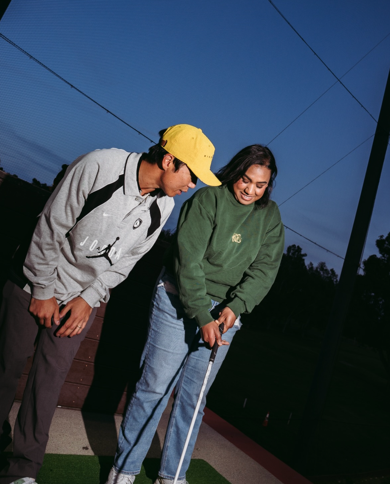 Two people standing outdoors at dusk, one holding a golf club, the other leaning in, with a blue sky and trees in the background.