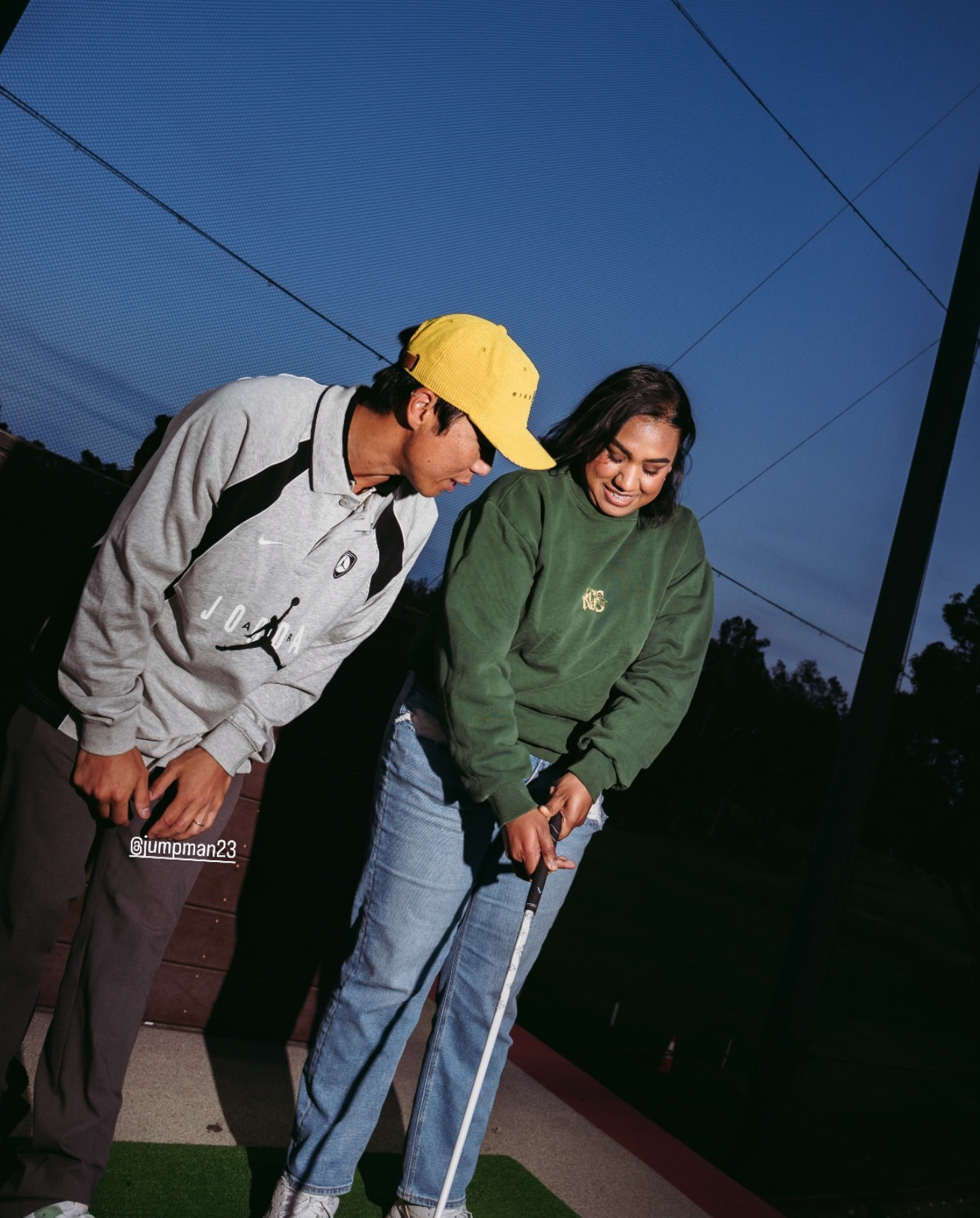 A young man and woman standing on a golf course at dusk, holding golf clubs and smiling. The man is wearing a gray sweatshirt with a Jumpman logo and a yellow cap. The woman is wearing a green sweatshirt and ripped jeans.