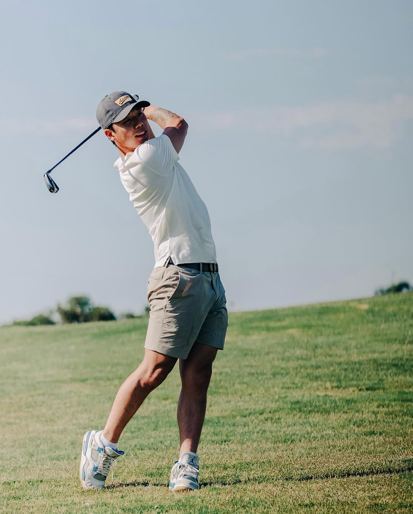 A man playing golf on a grassy course, swinging with a golf club, wearing a gray cap, white shirt, and khaki shorts.