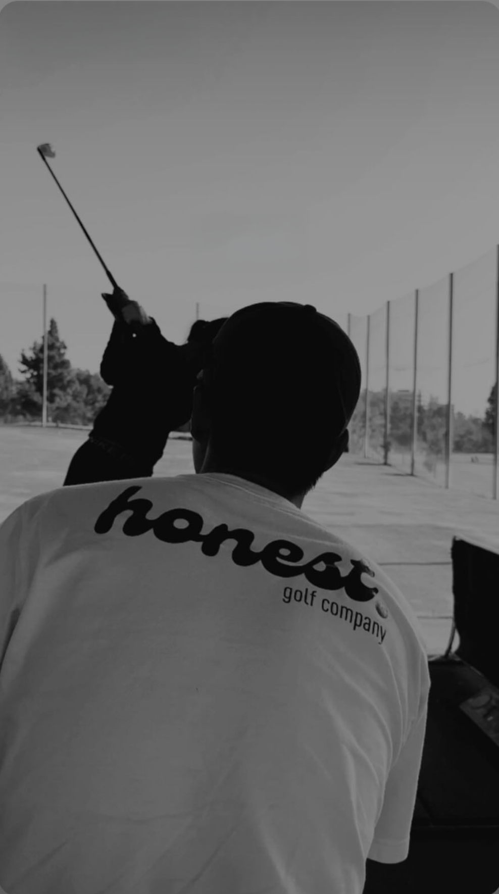 A person wearing a Nike Golf Company shirt is sitting near a golf driving range, while another person in a dark jacket swings a golf club in the background.