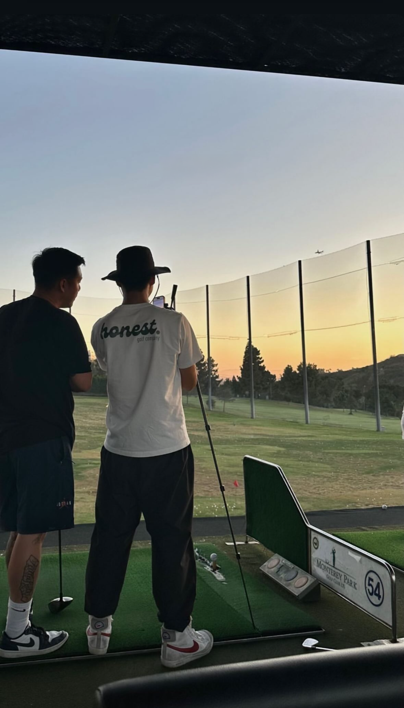 Two men standing at a driving range, one wearing a wide-brimmed hat and the other in sportswear, with a golf club, practicing golf during sunset. There are golf balls on the ground, a golf sign, and safety netting with trees and a plane flying overhead.